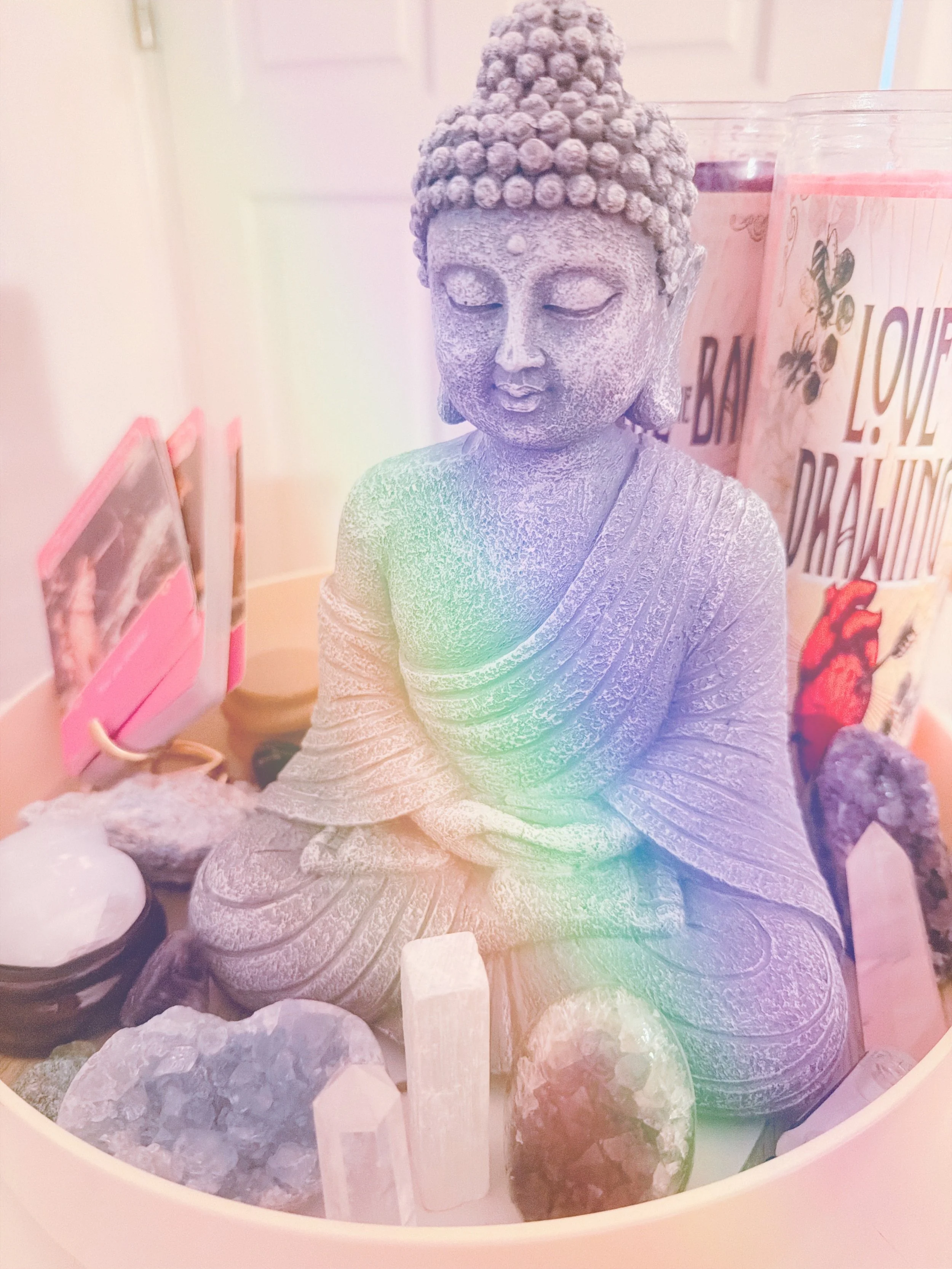 A gray statue of Buddha with closed eyes, seated cross-legged, surrounded by crystals and small objects, with pink candles and decorative items in the background.