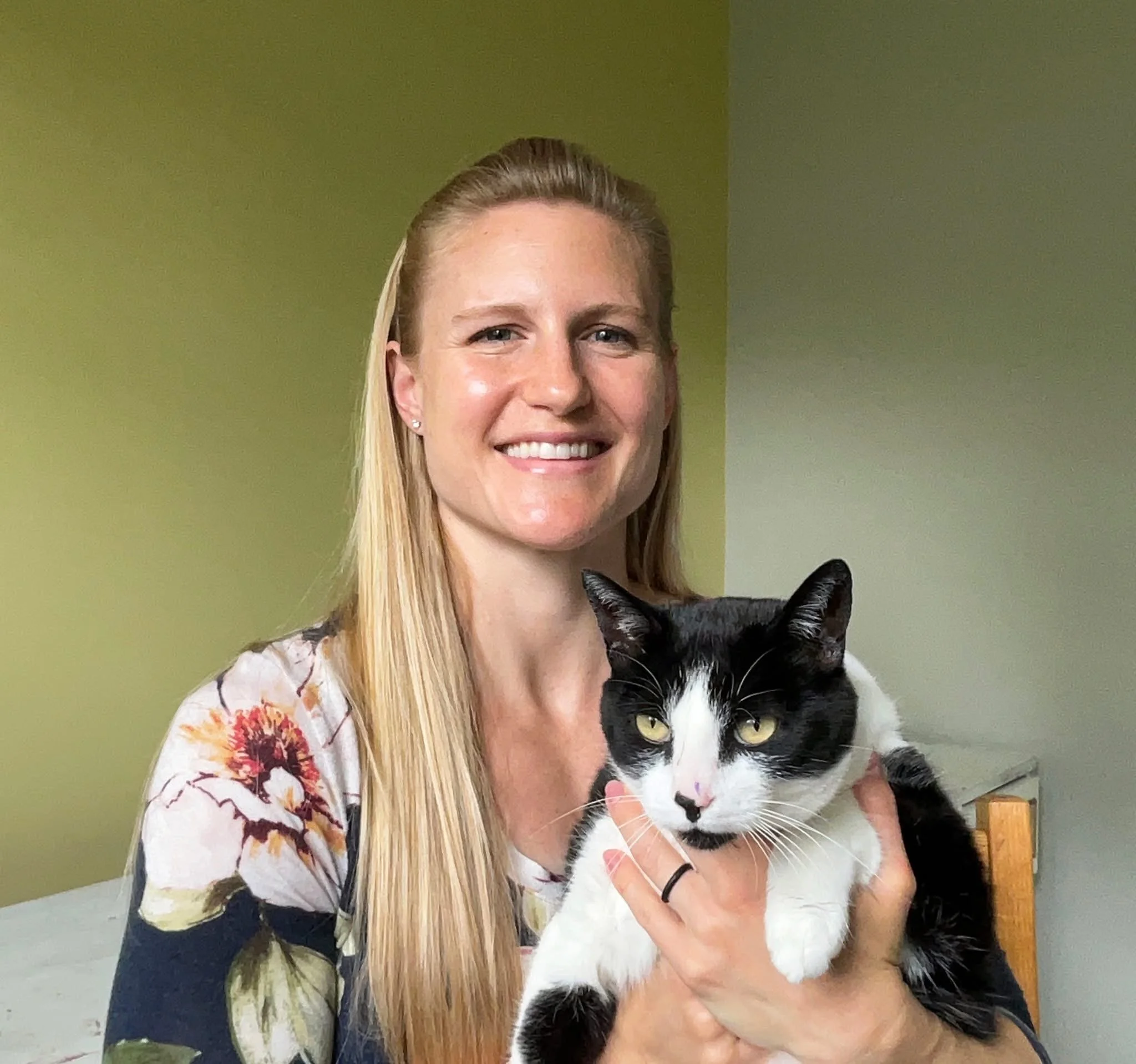 A woman with long blonde hair smiling and holding a black and white cat indoors with green walls.