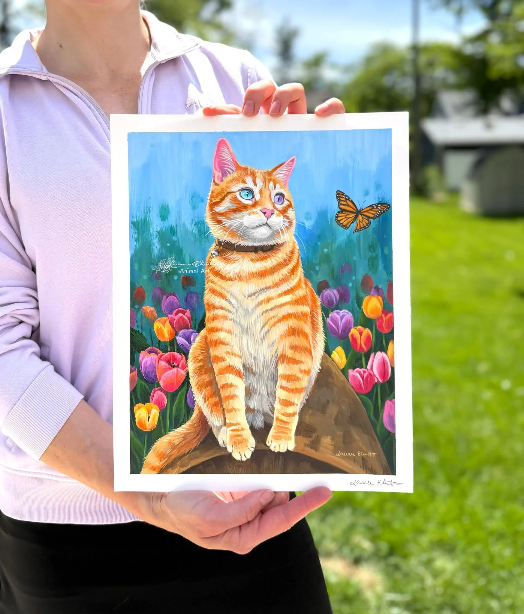 A person holding an illustration of a cat with a blue eye and a purple eye, surrounded by colorful tulips and a monarch butterfly, outdoors on a sunny day.