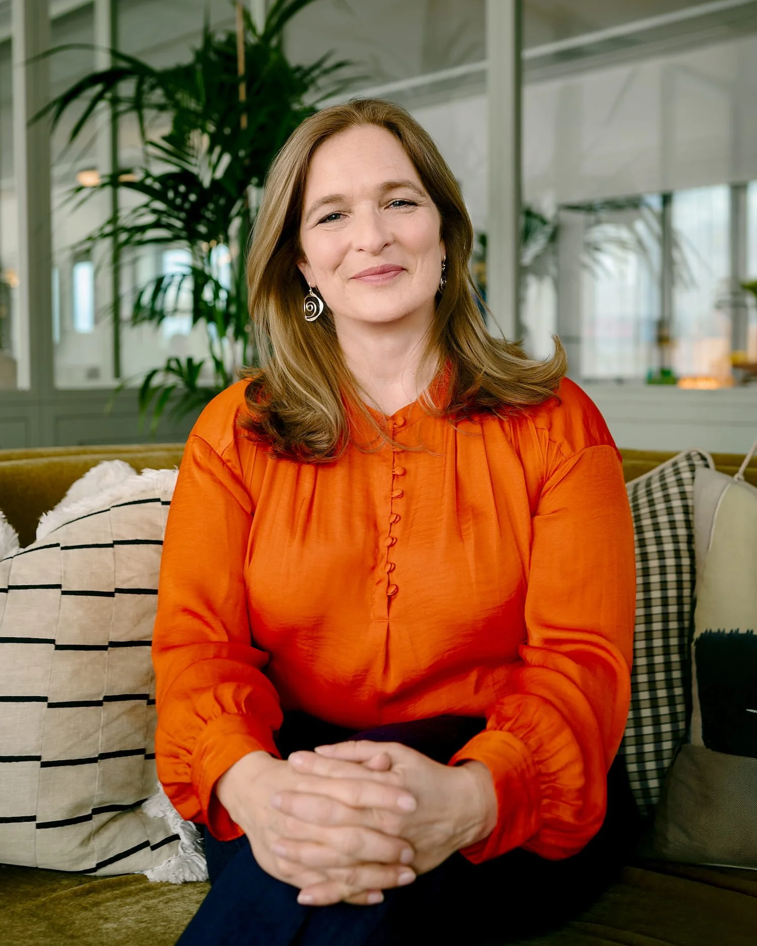 Mari Williams, Executive Therapist & Leadership Coach smiling on sofa with hands crossed, orange blouse