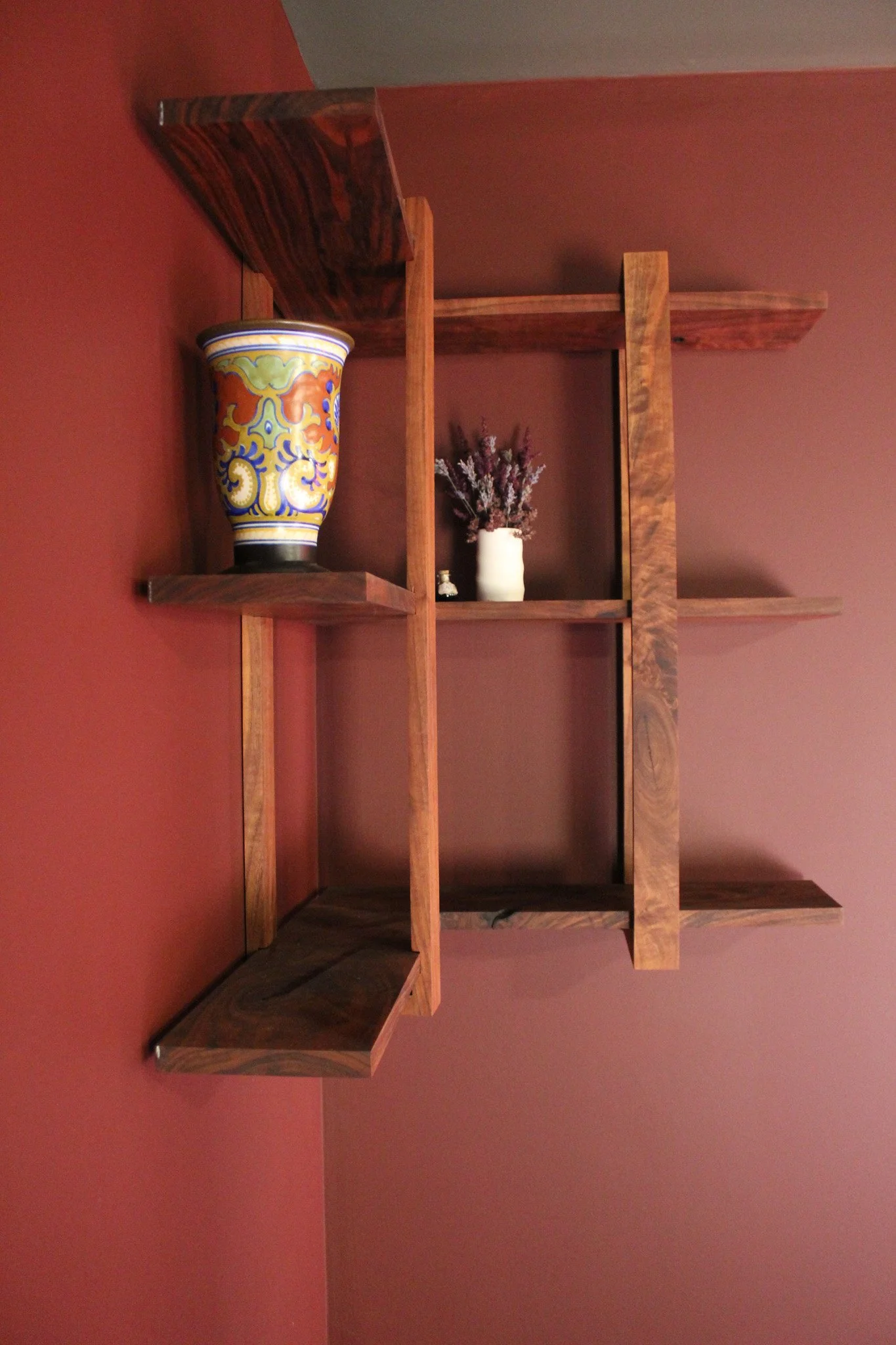 Wooden wall shelf with three sections, decorated with a colorful ceramic vase on the left, a white vase with dried flowers in the middle, and the empty right section, against a reddish wall.