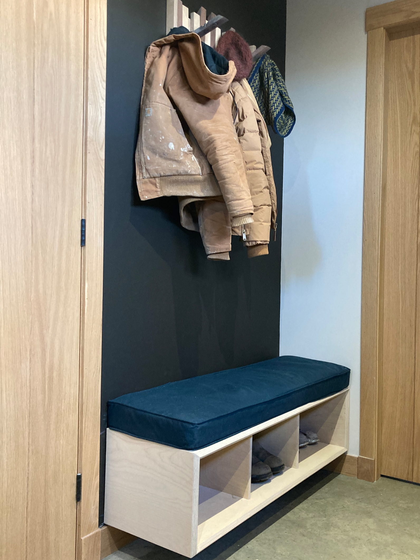 Coat rack with jackets, hats, and scarves above a wooden hall bench with storage cubbies and a cushioned seat in a hallway corner.