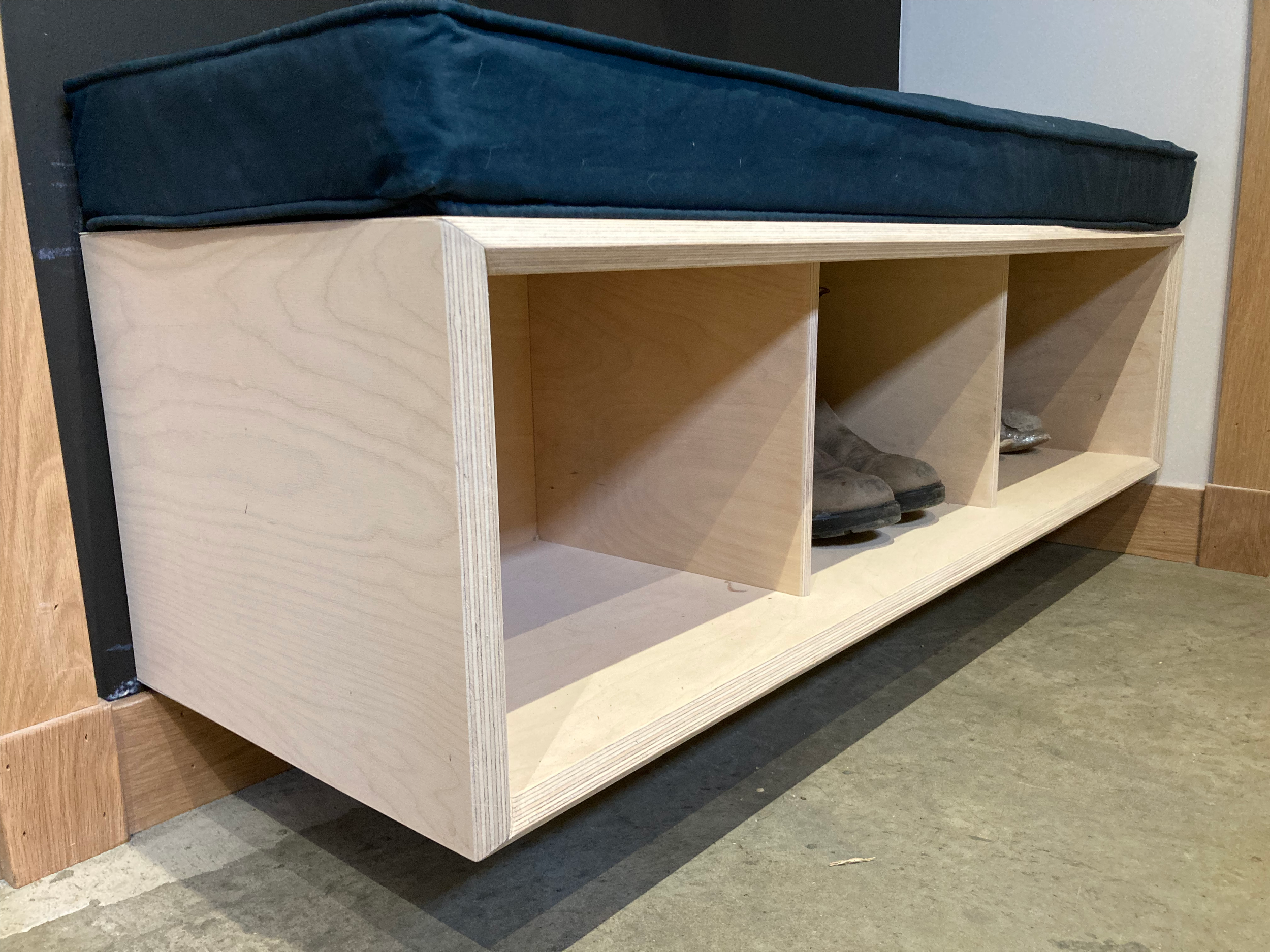 A wooden furniture piece with a cubed design, featuring three open shelves and shoes placed on the shelves, positioned on a floor with mixed wood and tile surfaces.