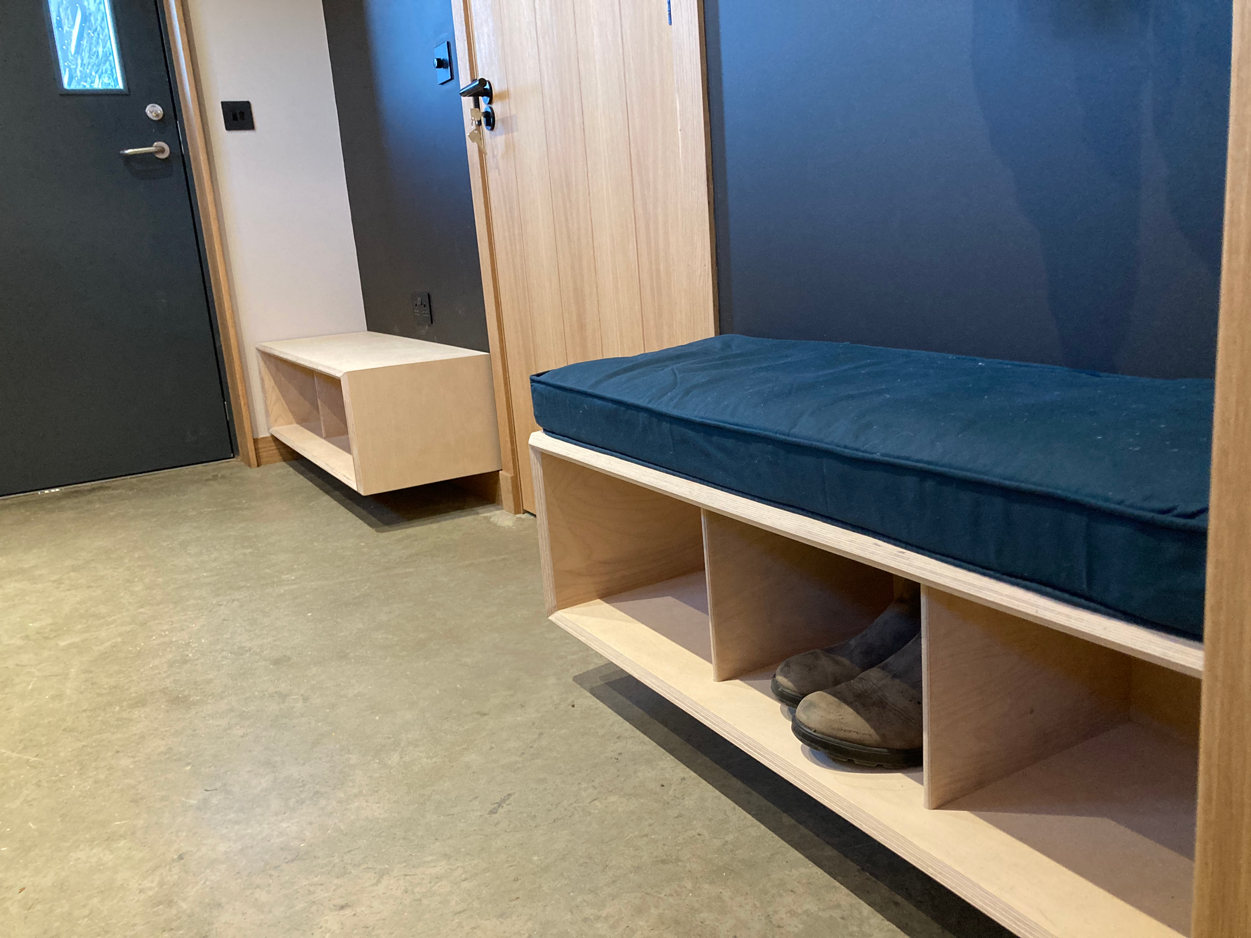 Photo of an interior space showing a wooden shelf with shoes, a blue cushioned bench, a door, and electrical outlets on the wall.