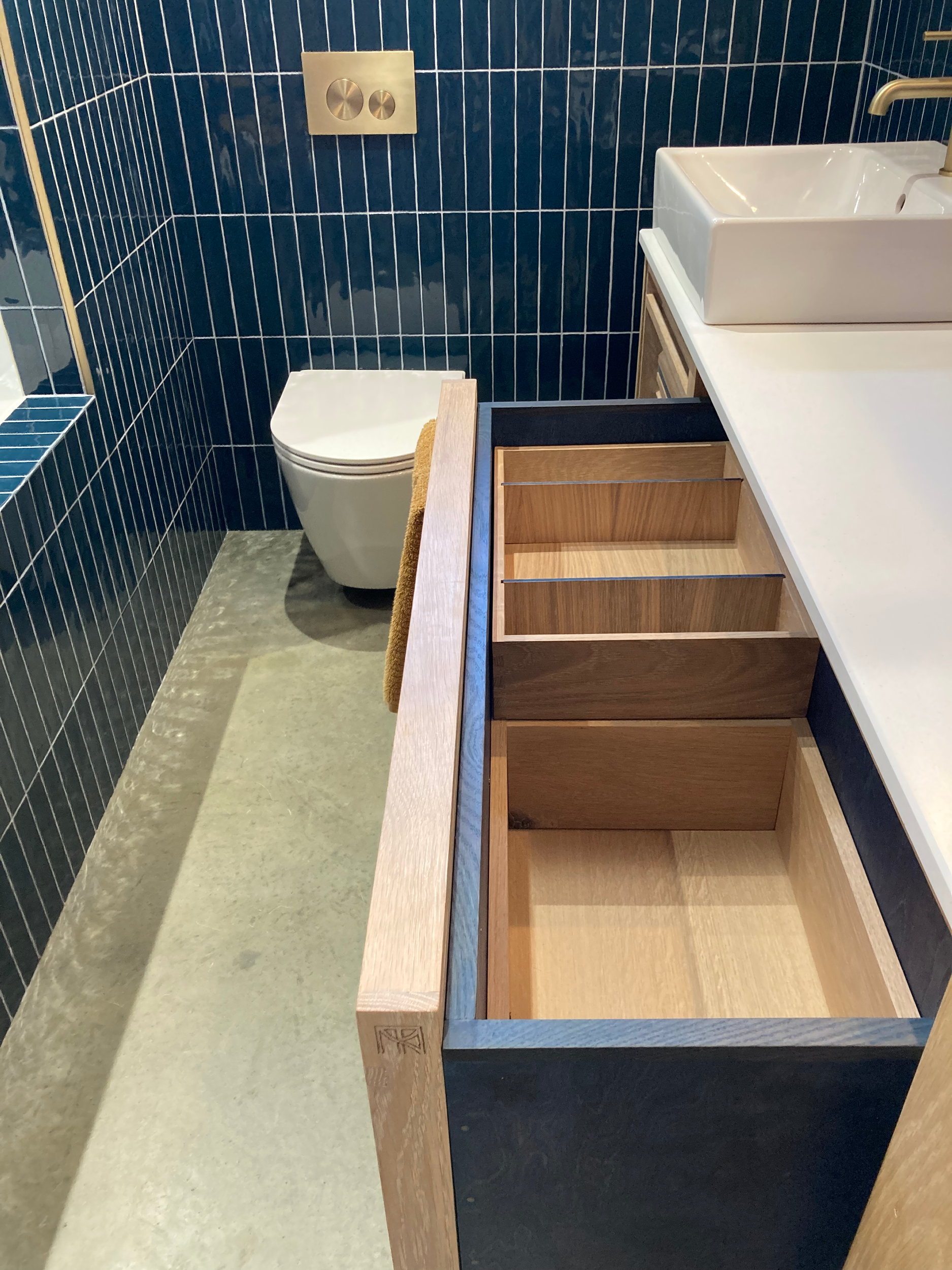 Modern bathroom with a dark blue tiled wall, a white sink, and a wooden storage shelf. A section of a toilet is visible.