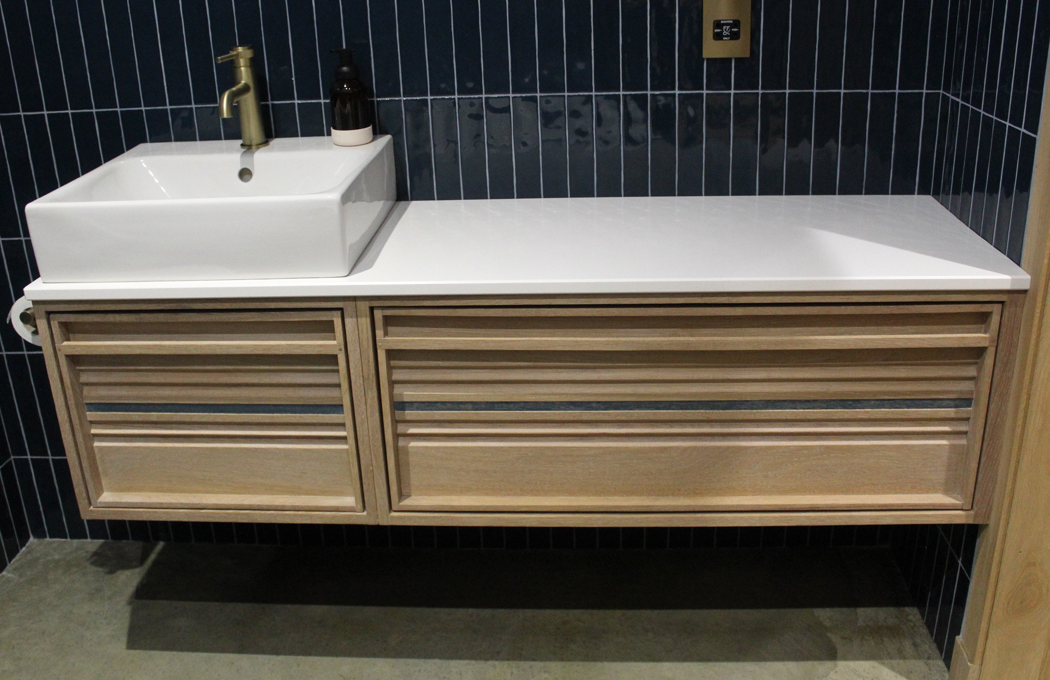 Modern bathroom vanity with a white rectangular vessel sink, brass faucet, black soap dispenser, wooden cabinet with slatted doors, black tiled wall, and concrete floor.