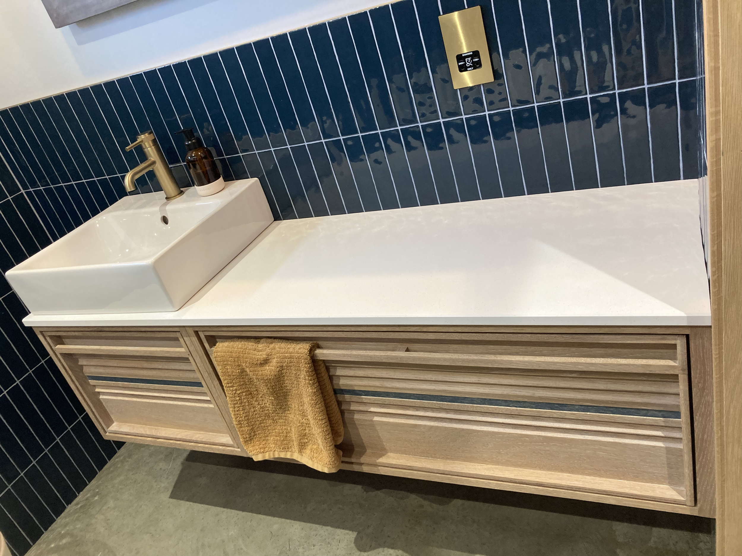 Modern bathroom vanity with a rectangular white vessel sink, gold faucet, amber soap dispenser, yellow hand towel, and blue tiled wall with a gold outlet.