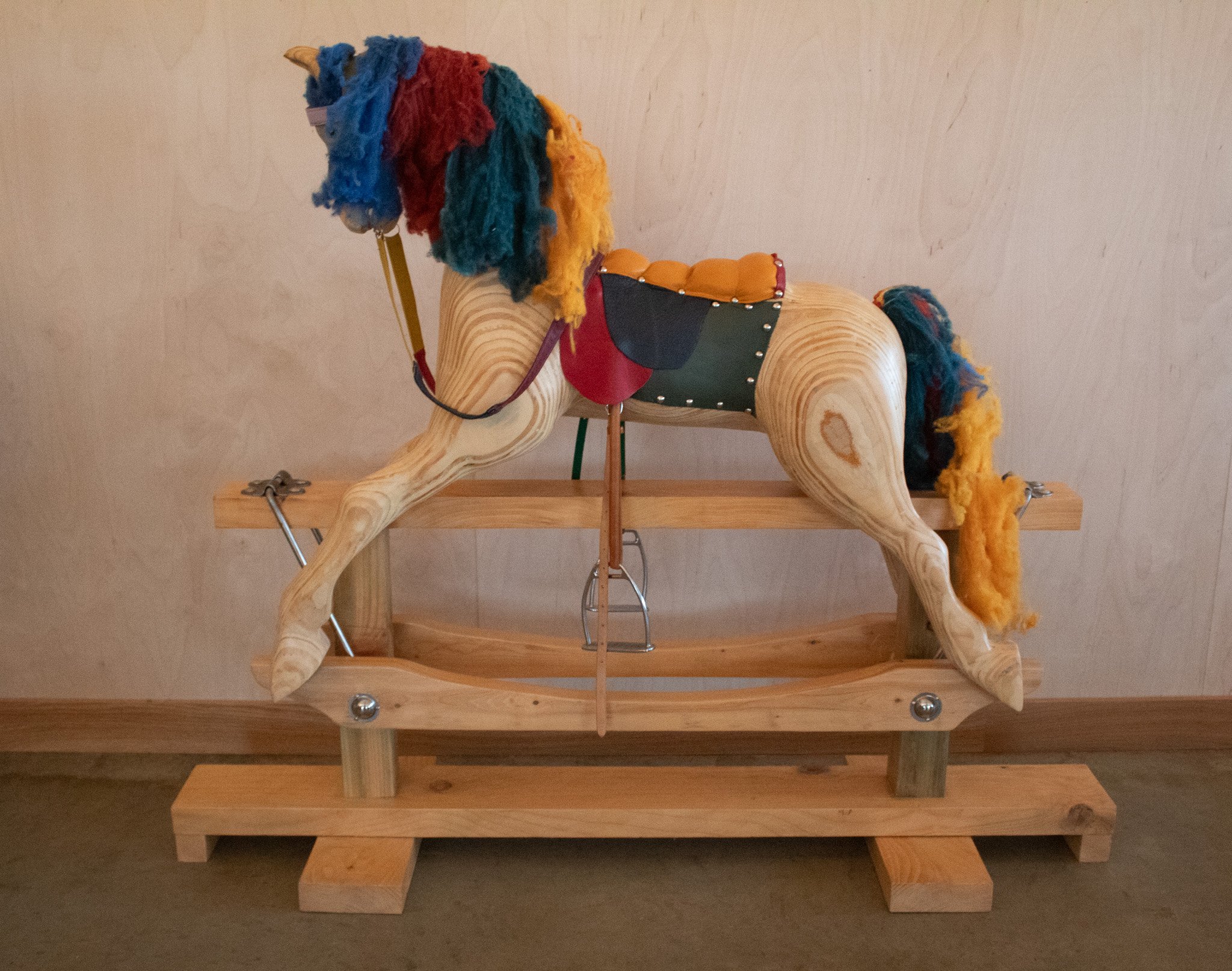 A wooden rocking horse with a colorful mane made of multicolored yarn, placed on a wooden stand against a beige wall.
