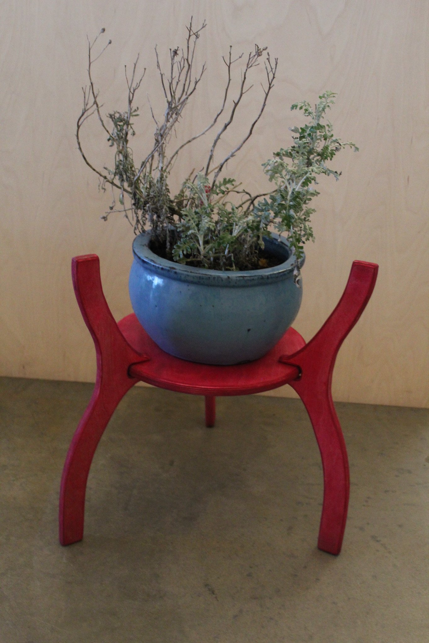 A blue ceramic pot with dried and green plants sits on a distinctive red wooden stand with three curved legs, against a plain beige wall.