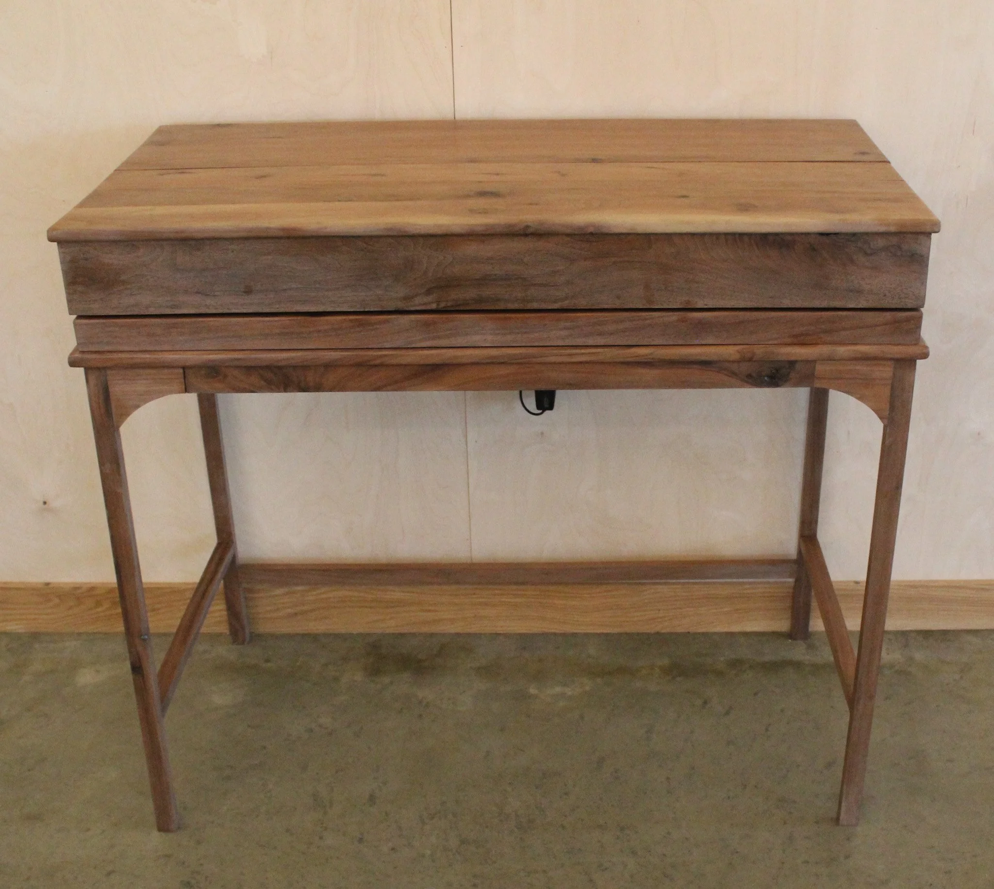 Wooden table with a closed lid on top and a lower shelf, standing on a concrete floor against a light-colored wall.