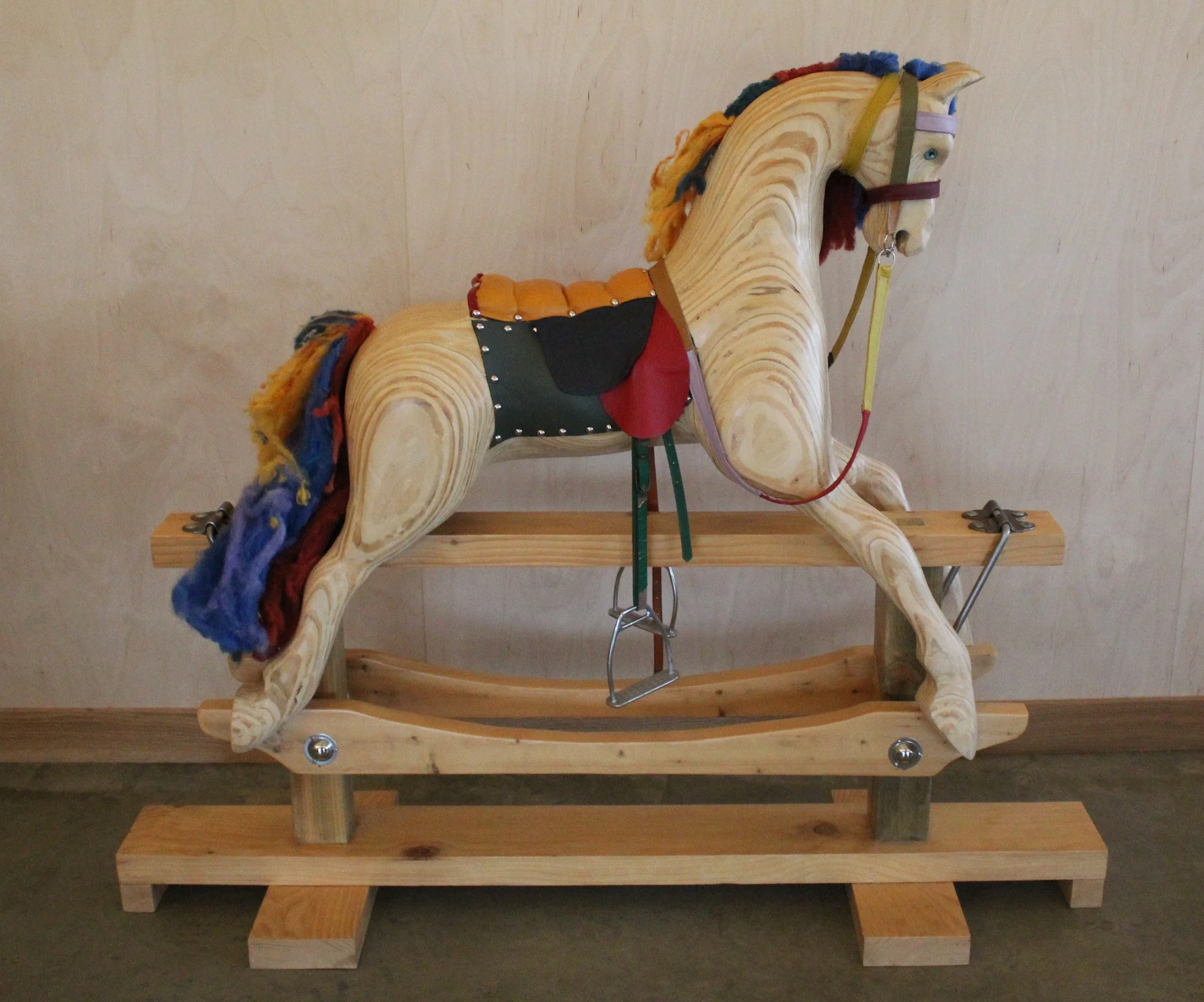 Wooden rocking horse with colorful mane and tail, saddle, bridle, and stirrups, mounted on a wooden base.