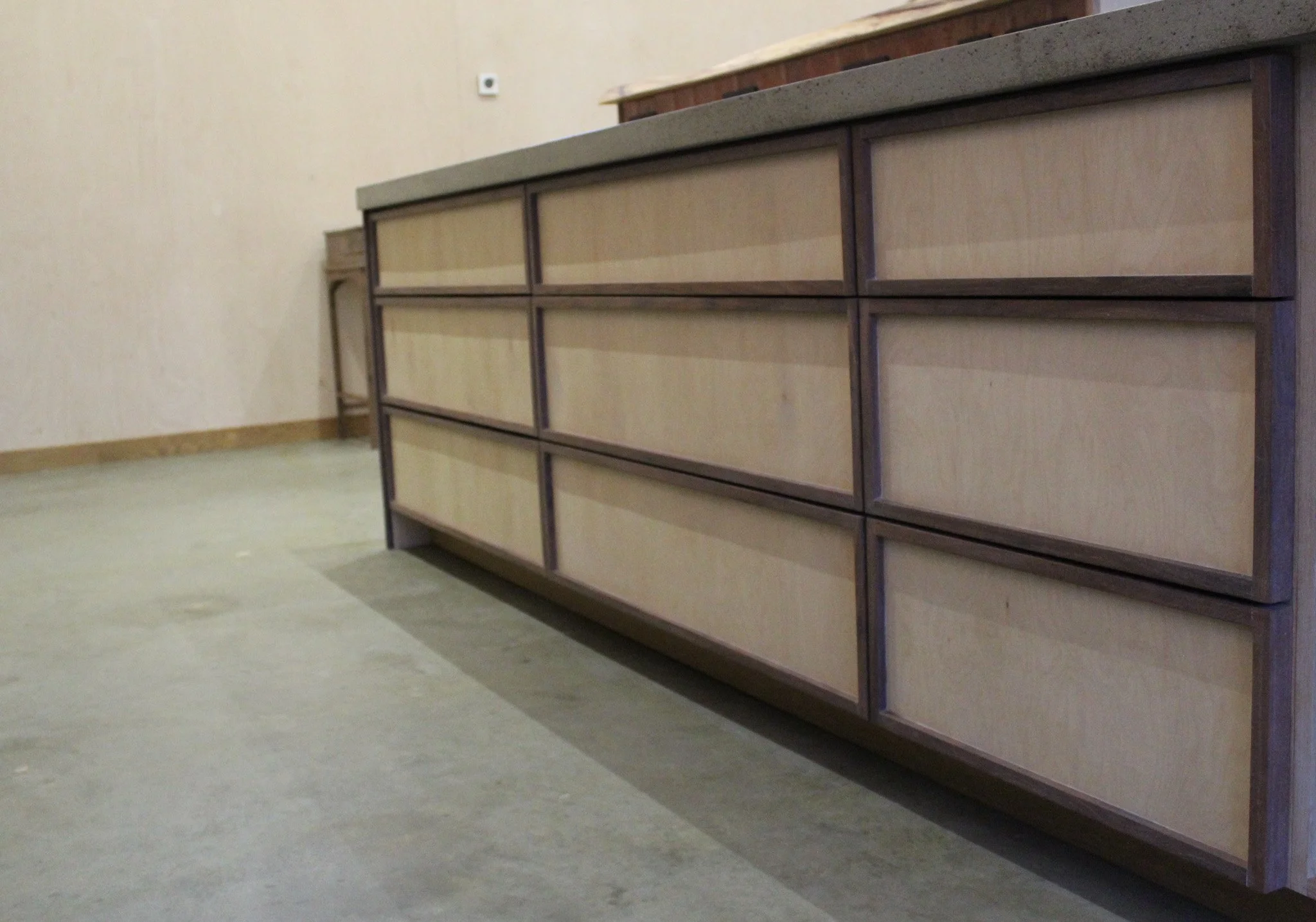 Empty wooden cabinet with multiple drawers in a room with beige walls and a concrete floor.