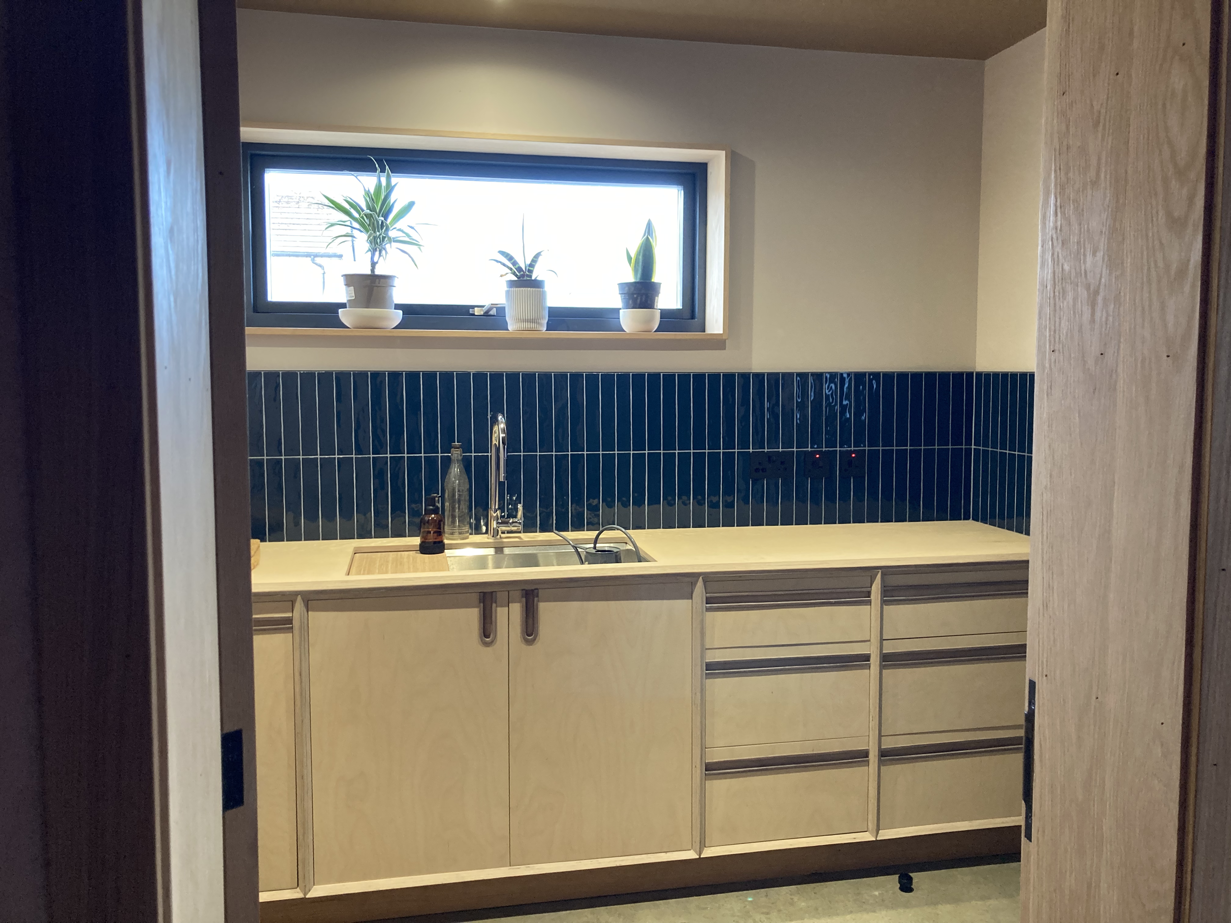 A modern kitchen with a beige wooden cabinet, black tile backsplash, and a window with three potted plants.