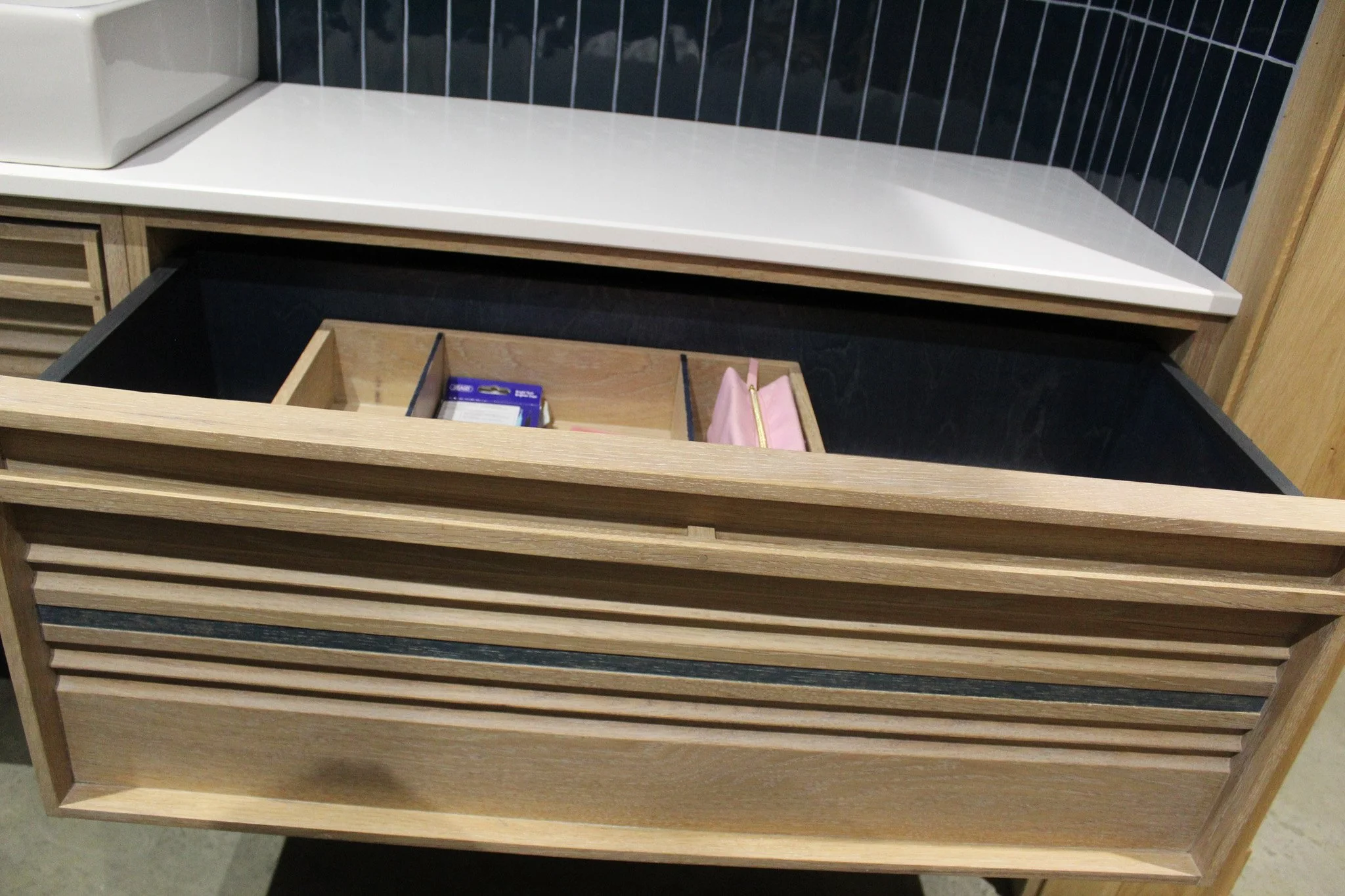 Open wooden drawer with black interior, containing a small pink pouch with a gold zipper and a blue pack of batteries, in a modern kitchen with white countertop and black tile backsplash.