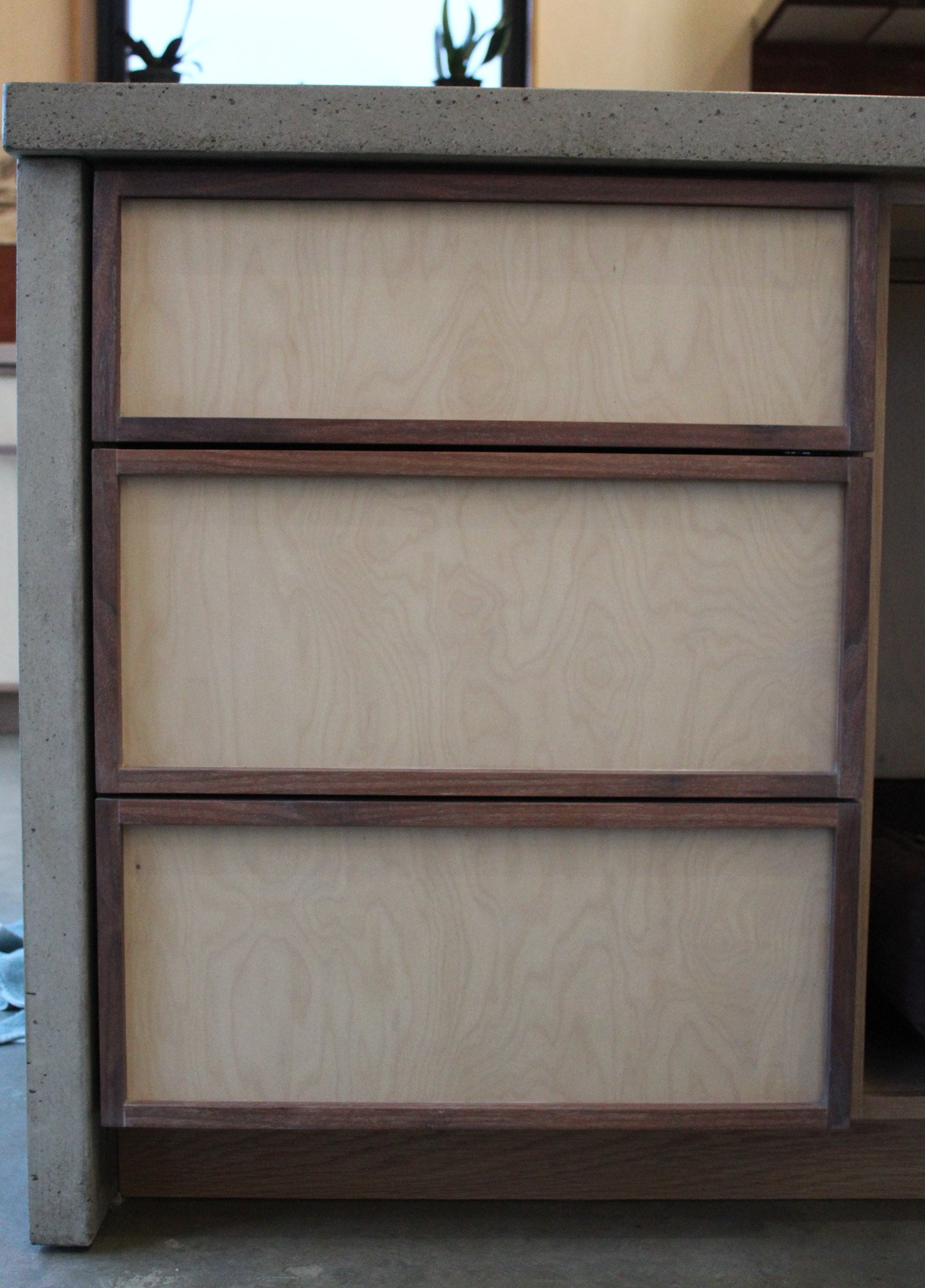 Close-up of a three-drawer wooden cabinet with light-colored drawer fronts and darker wooden edges, set against a stone countertop.