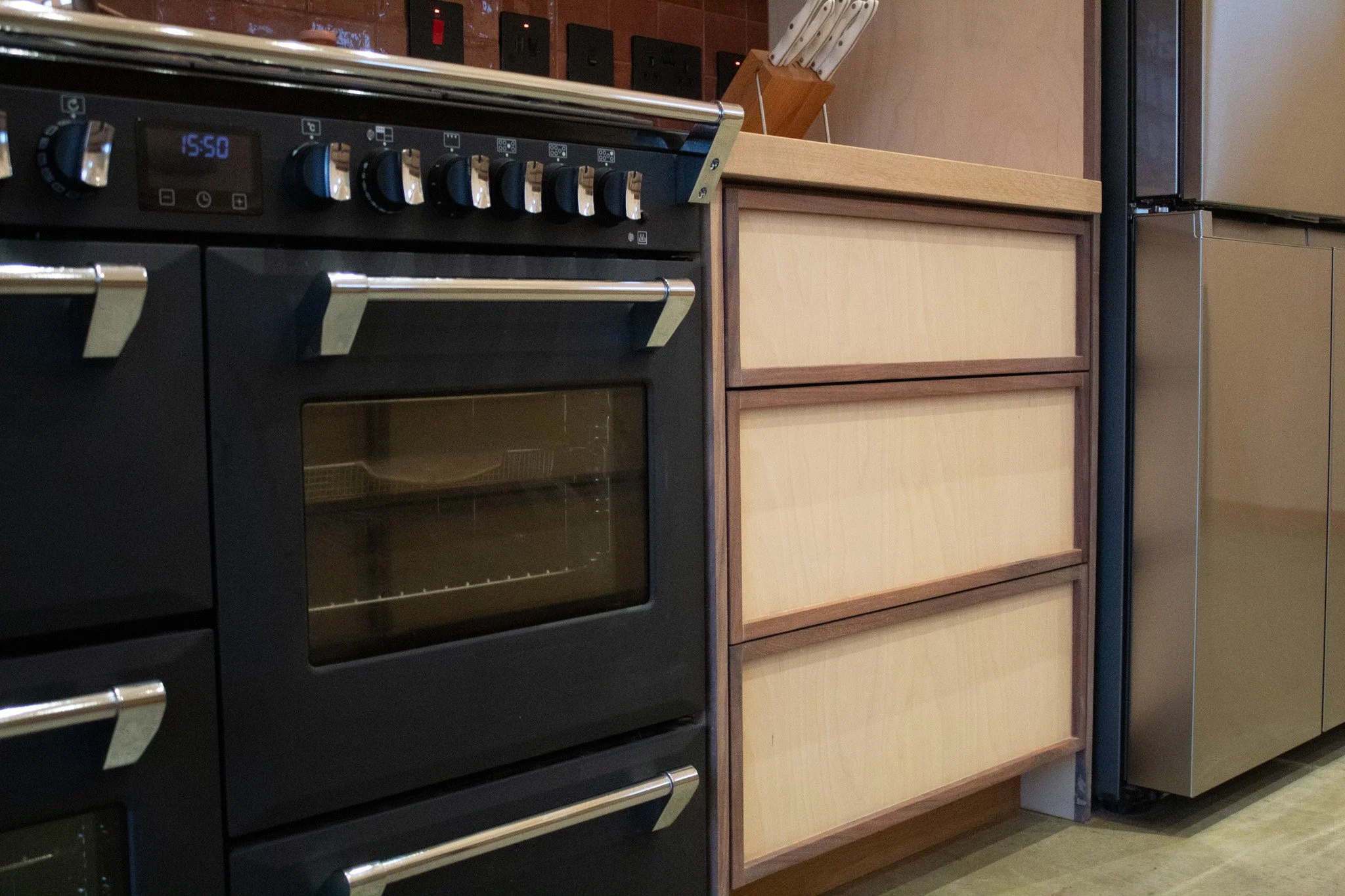 A black oven with digital clock and control knobs, wooden drawers to the right, and silver refrigerator on the far right in a kitchen.