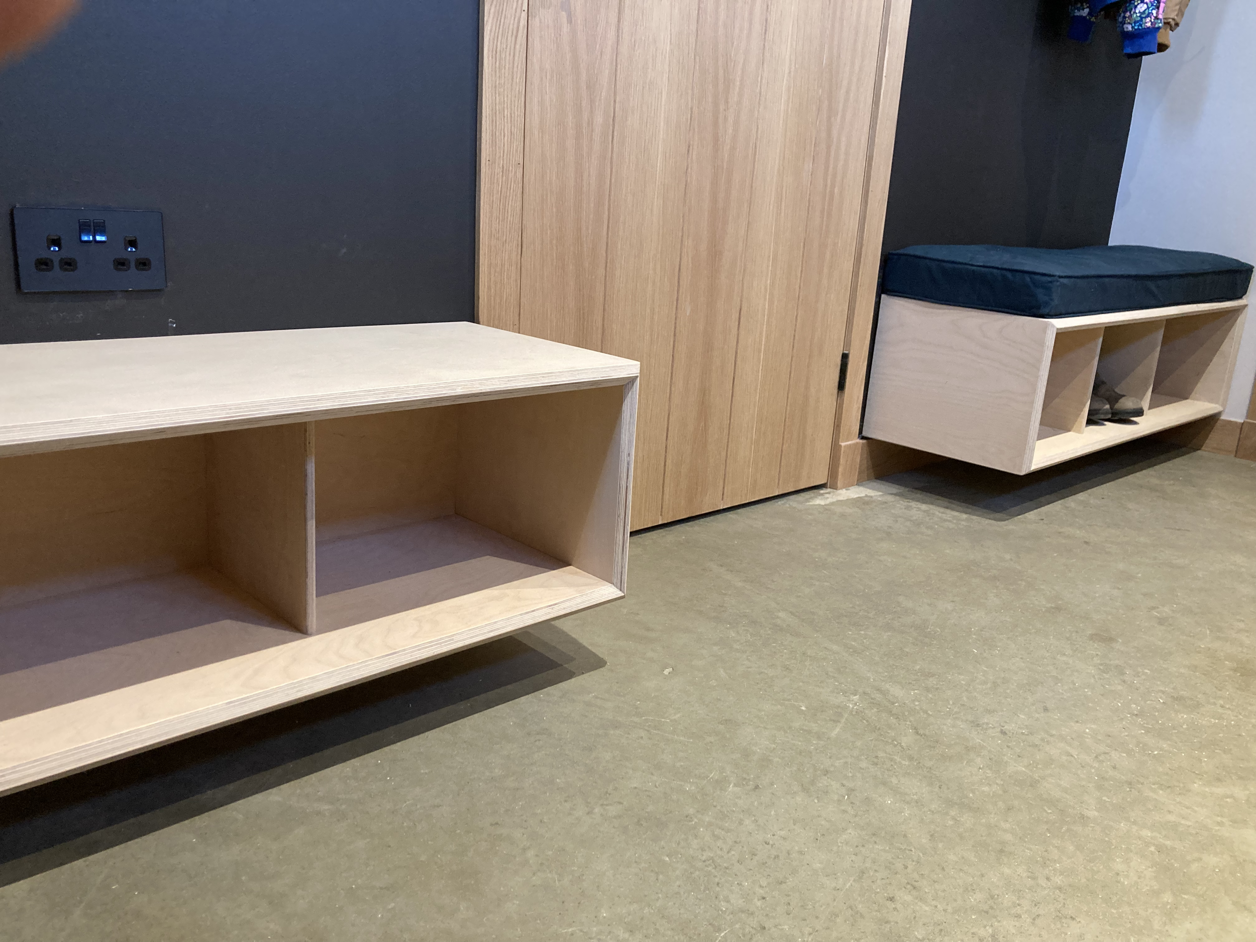 Interior of a room featuring two light wooden benches with open cubbies underneath, one with a dark cushion and the other empty, against a wall with a dual power outlet.