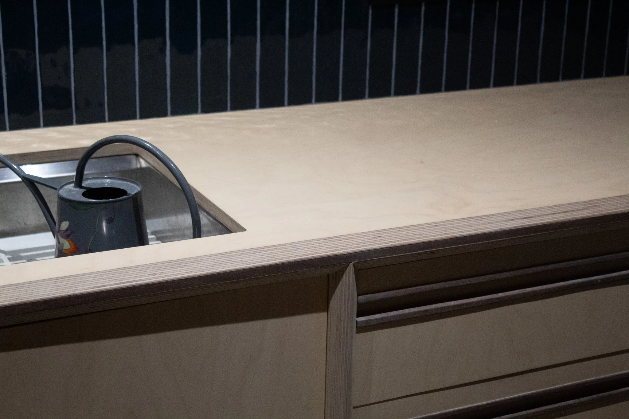 A close-up of a wooden countertop with a stainless steel sink in the corner. There is a black kettle with a floral design inside the sink. The background is dark with vertical railing bars.