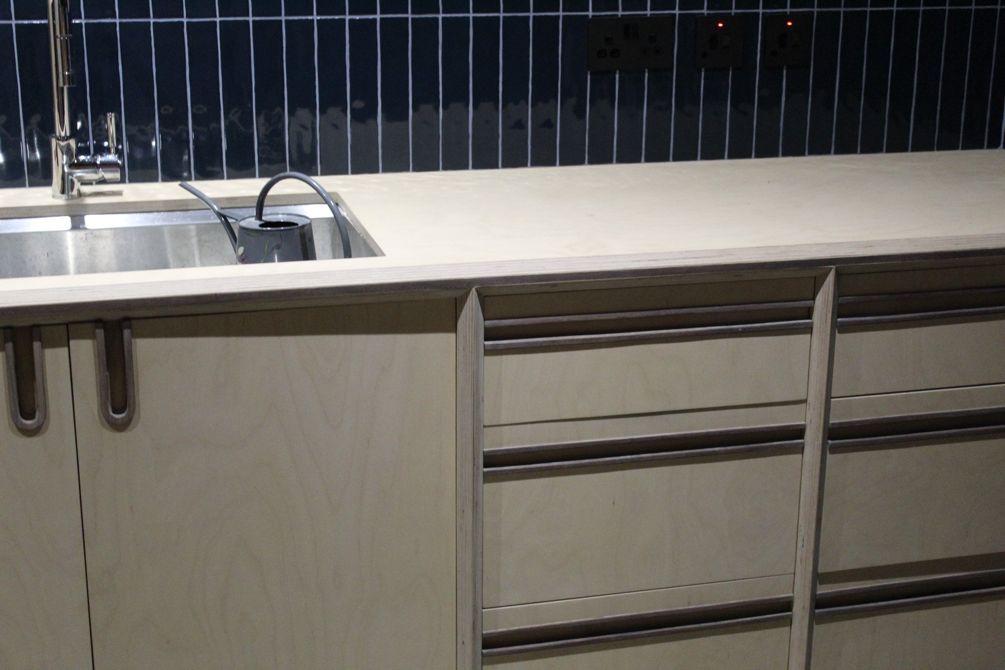 Kitchen countertop with a sink and a KitchenAid electric kettle, dark blue tile backsplash, and light wood cabinetry with horizontal handles.