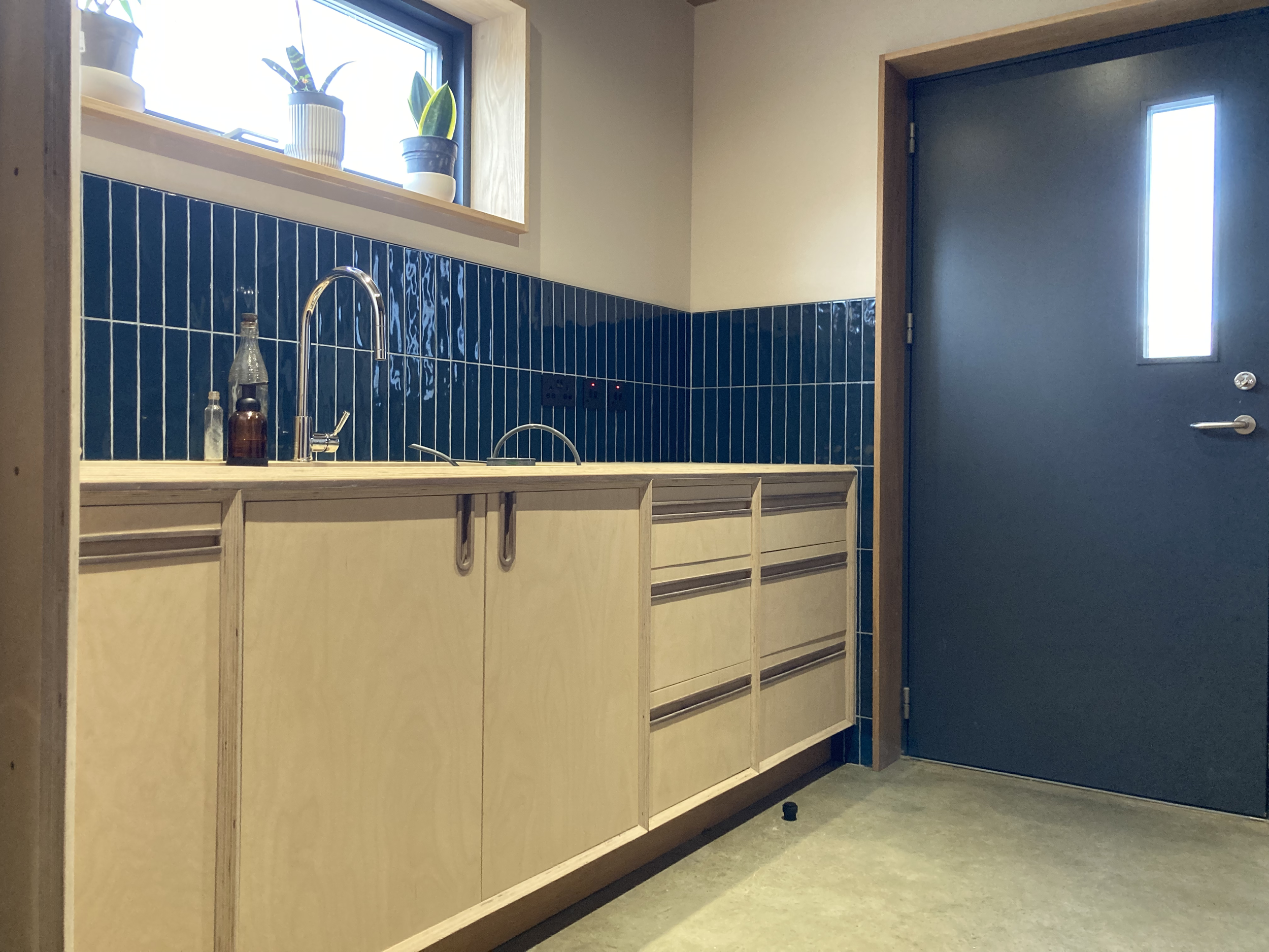 Minimalist kitchen with light wooden cabinets, blue tiled backsplash, two potted plants on the windowsill, a silver faucet, and a blue door with a window.