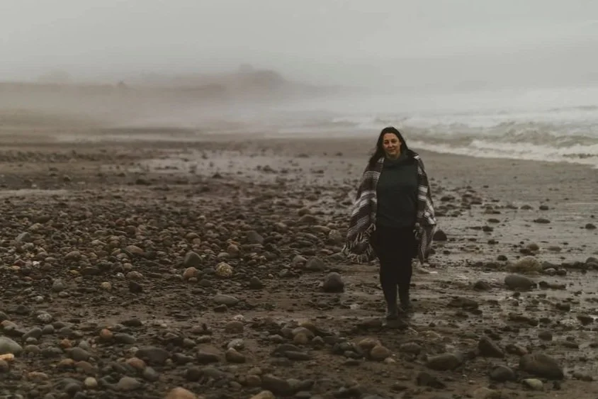 A woman standing on a rocky beach with the ocean and stormy sky in the background, wearing a striped shawl and black clothing.
