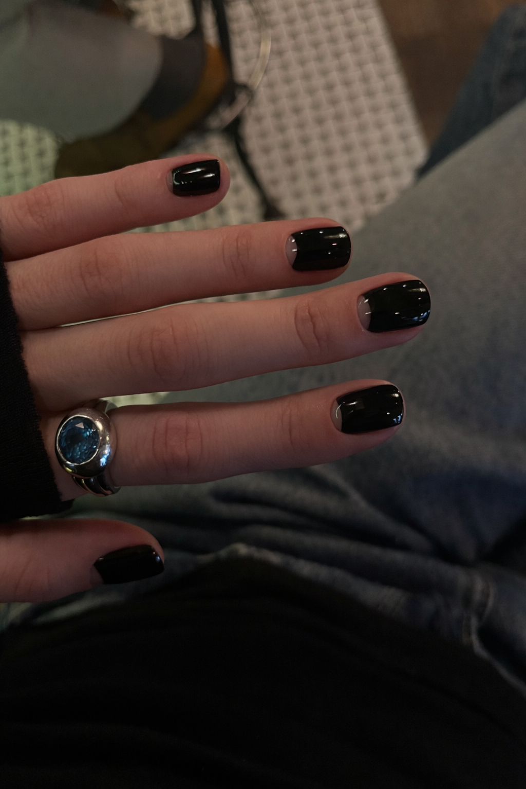 Close-up of a person's left hand with black painted nails, wearing a silver ring with a large blue stone and a black sleeve, resting on their lap.