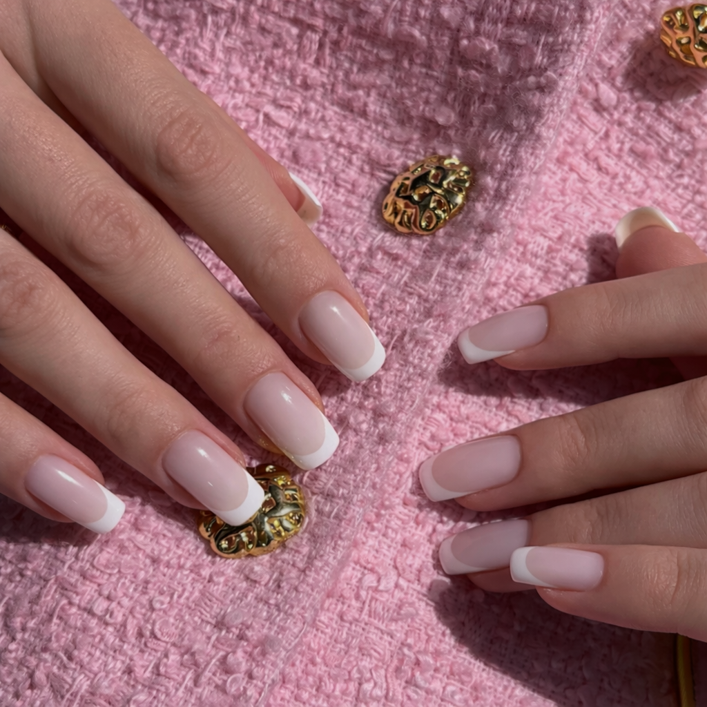 Close-up of hands showing manicured nails with French tips on pink textured fabric with gold decorative buttons.