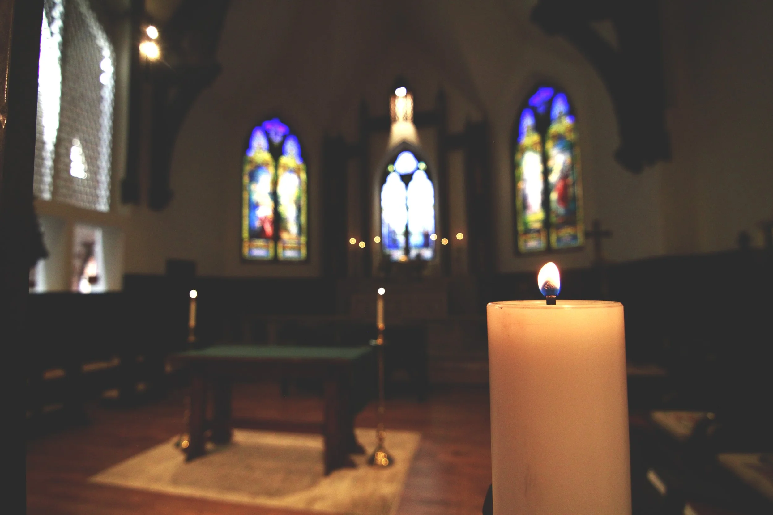 A lit candle inside a church with stained glass windows in the background.