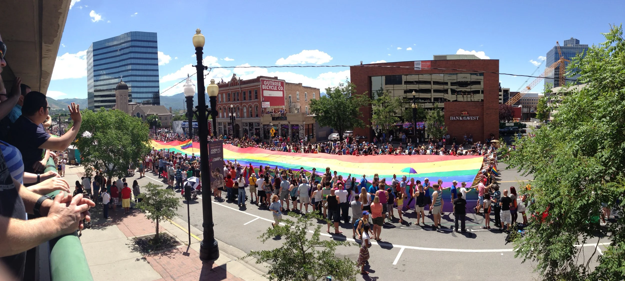 People holding a large rainbow pride flag during a parade in an urban area on a sunny day.
