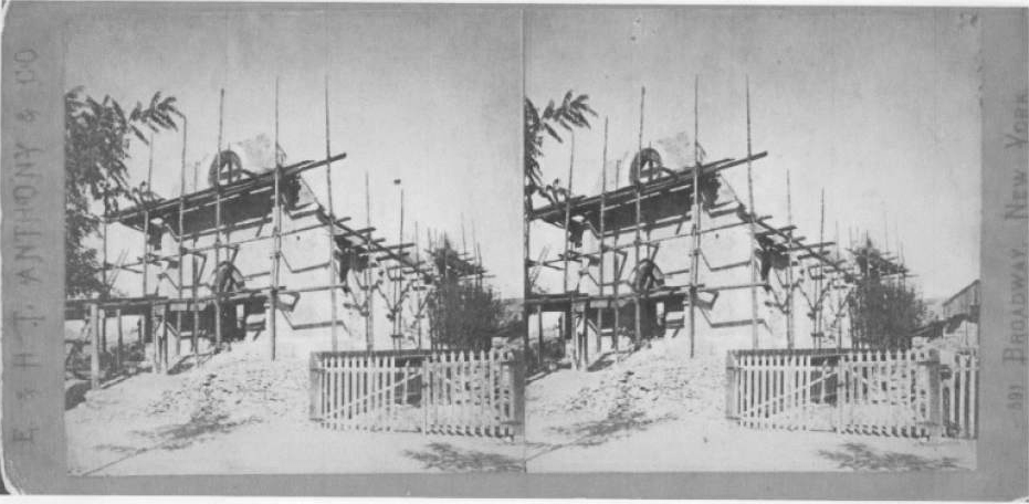 Black and white stereoscopic photograph of St Mark's Cathedral under construction, with scaffolding around it, a wooden picket fence in front, and trees nearby.