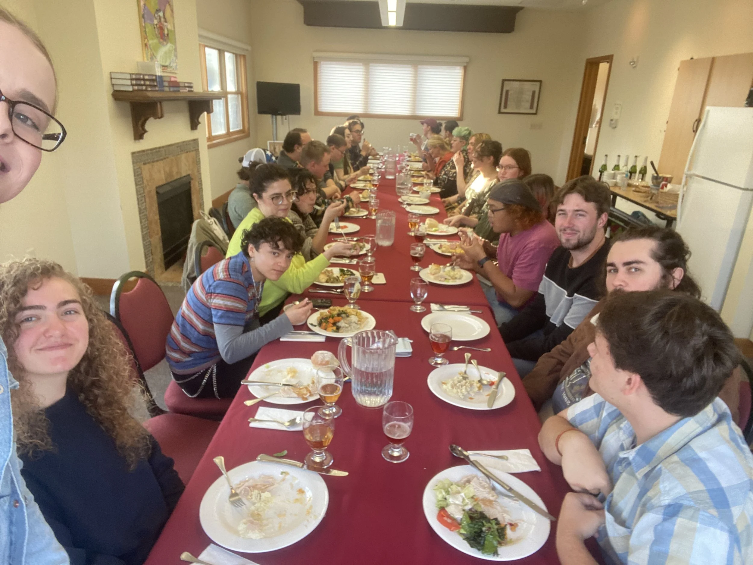 A large group of people enjoying a meal together at a long banquet table covered with a red tablecloth, in a room with windows, a fireplace, and a kitchen area in the background.