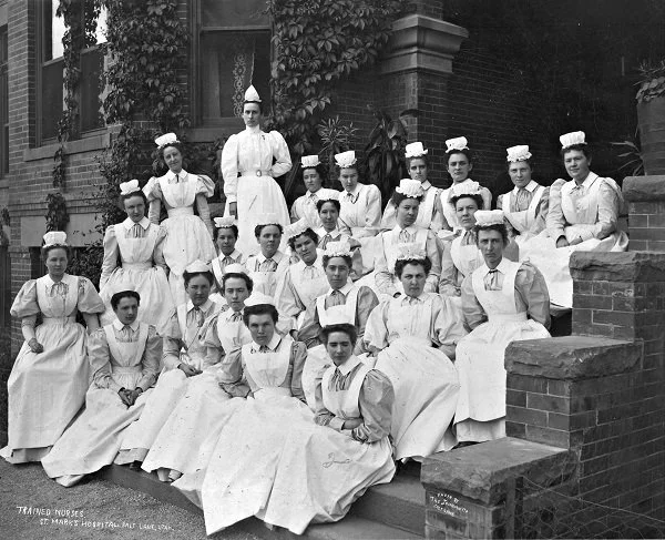 A group of nurses in white uniforms and caps sitting and standing on steps outside a brick building.