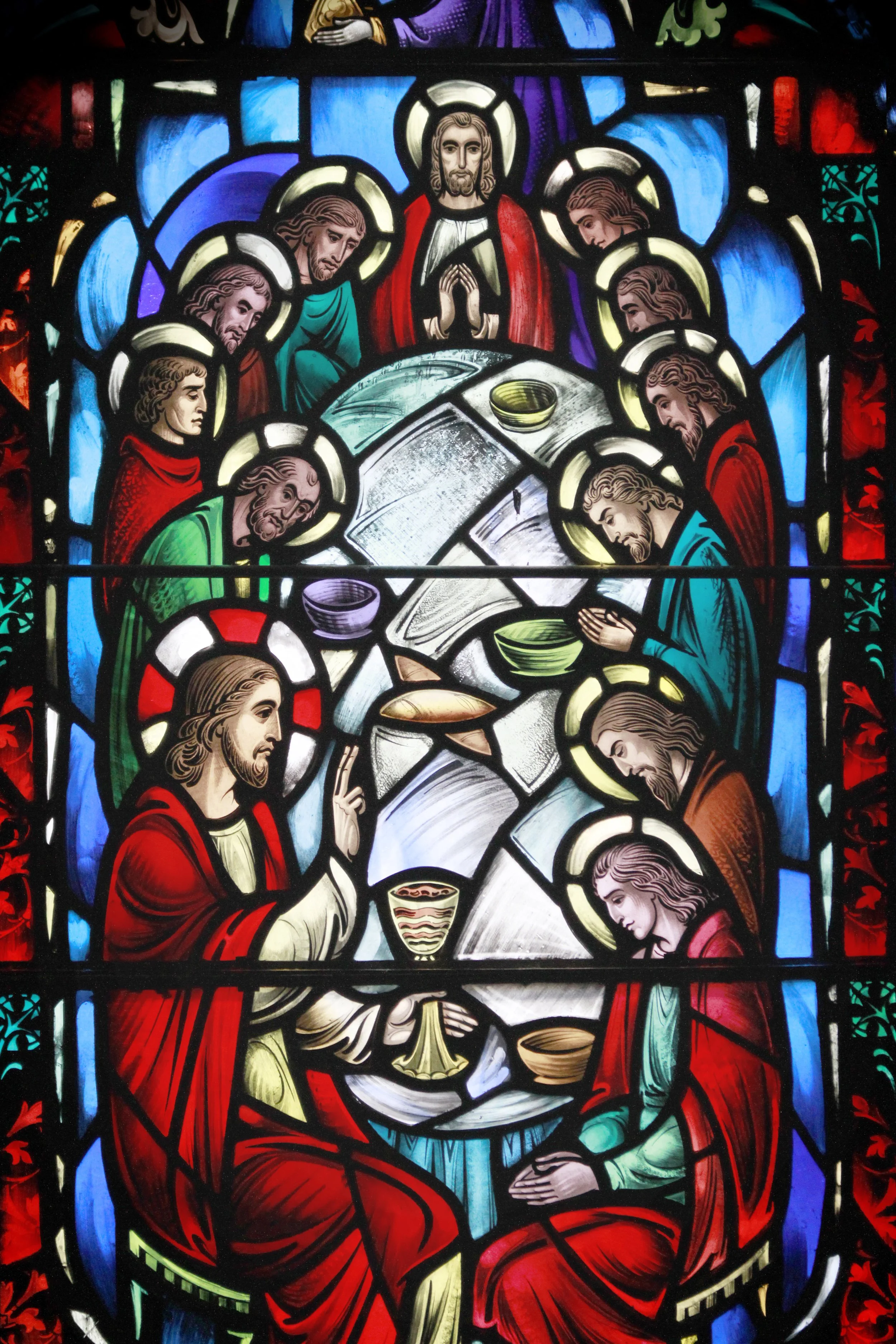 A stained glass window depicting Jesus Christ and his disciples during the Last Supper, with Jesus in a red robe at the center, surrounded by apostles with halos, seated around a table with bread and wine.