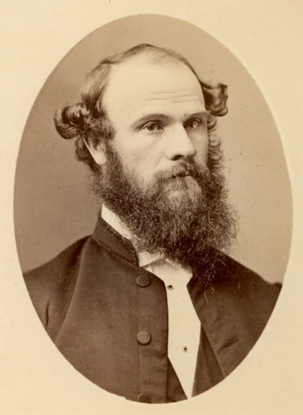 Black and white portrait of a man with a beard and mustache, wearing a suit with a high collar and bow tie, with hair parted in the middle and styled outward on the sides.