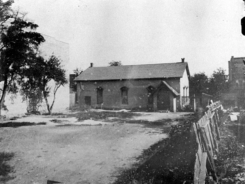 A black and white photo of early church with a gabled roof, surrounded by trees and a dirt yard, with a wooden fence on the right side.