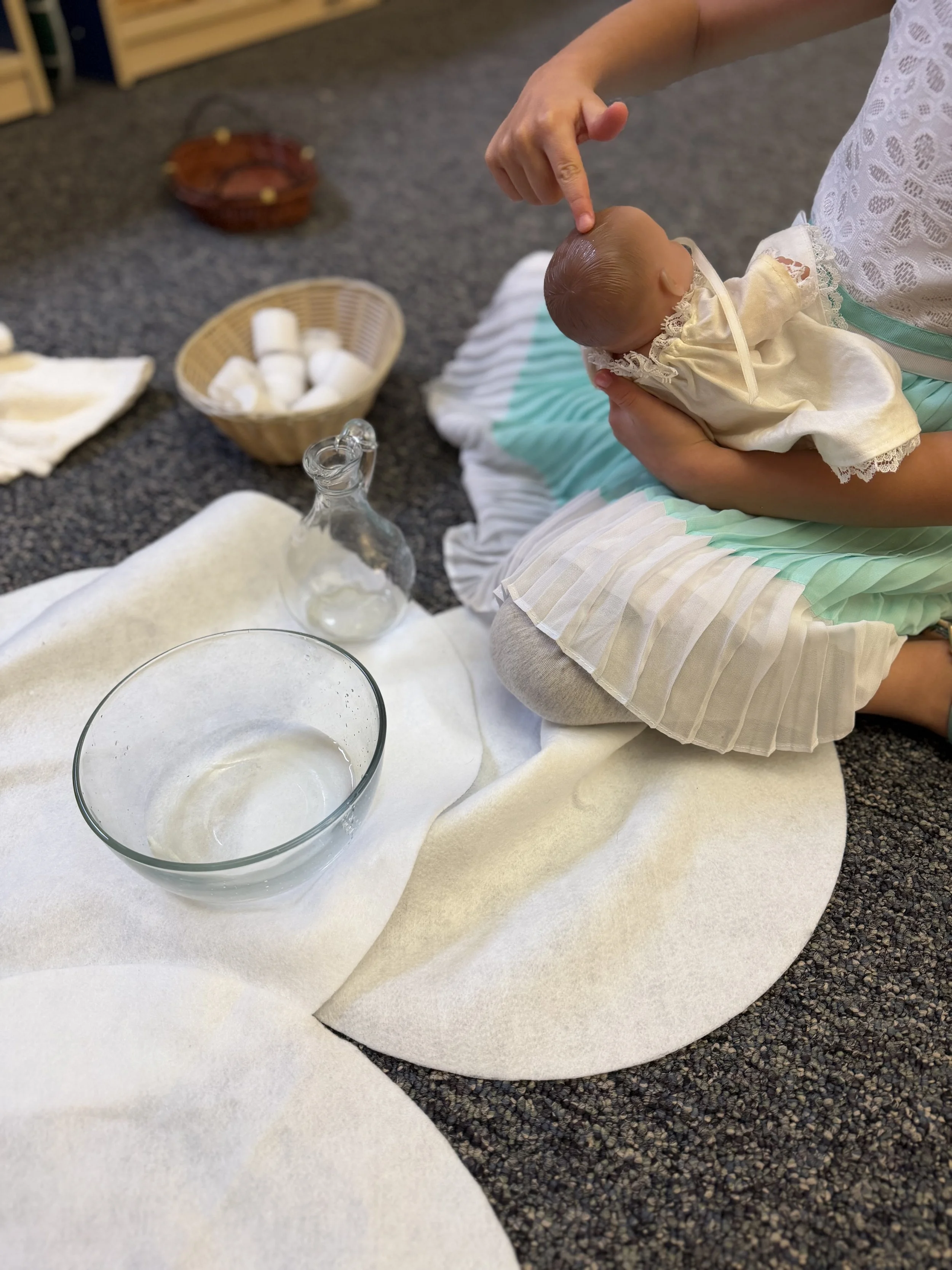A woman is holding a small baby, who is dressed in a light-colored dress, in an indoor setting with a carpeted floor. There are various items on the floor, including a bowl, a small glass bottle, a basket of rolled-up cloths or towels, and some white cups or containers.