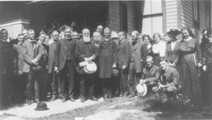 Group of men and women standing outdoors in front of a building, dressed in early 20th-century clothing, some holding hats.