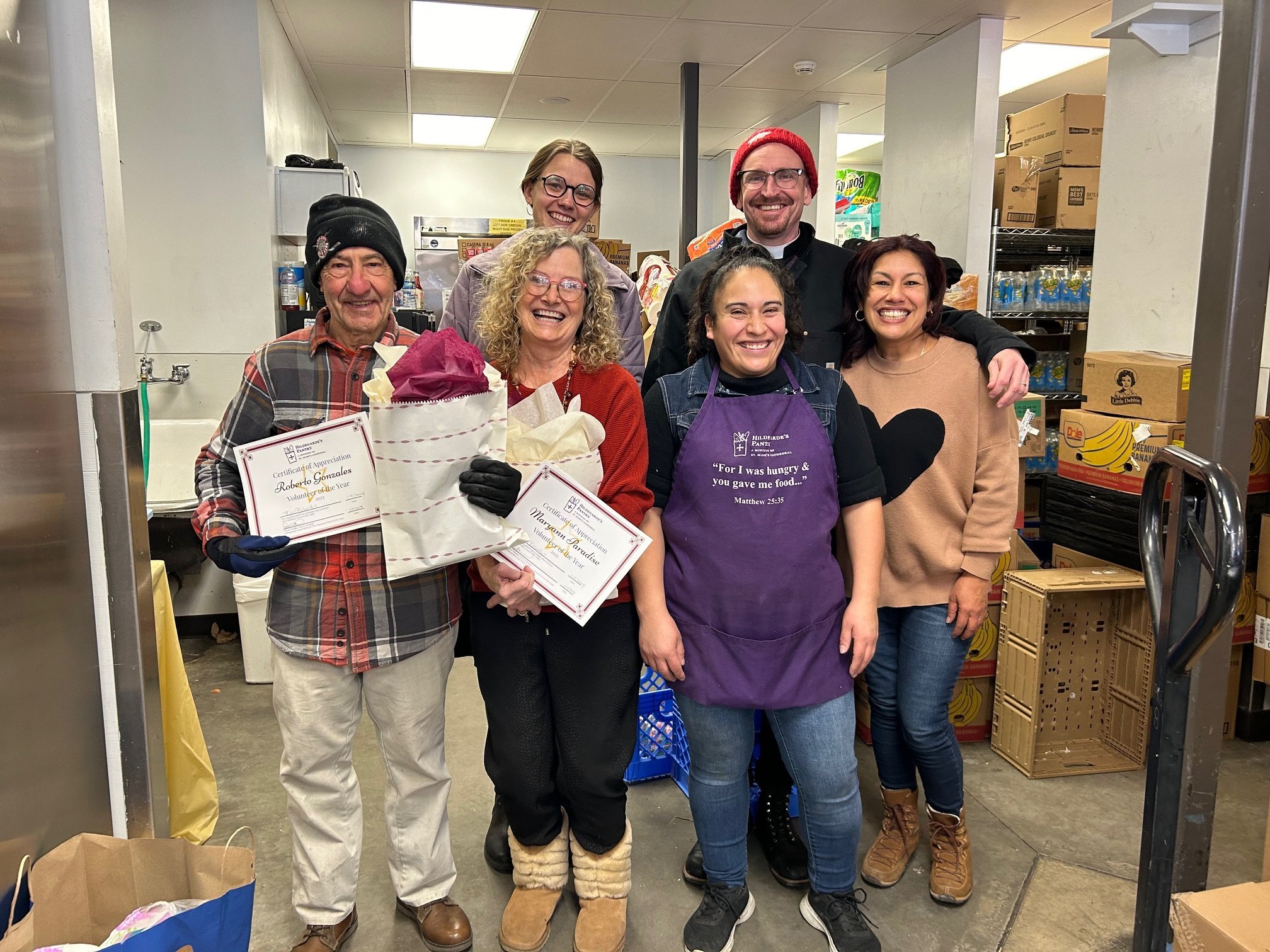 Group of seven volunteers smiling inside a food pantry, some holding certificates and gifts, surrounded by shelves of food items.