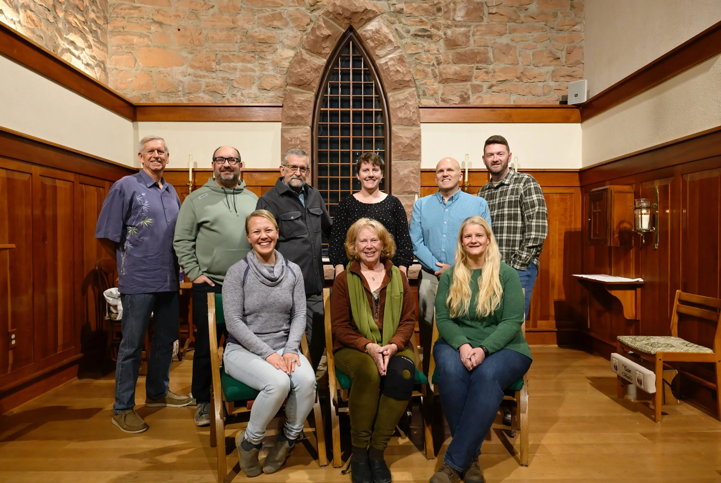 Group photo of ten people, five seated and five standing, inside a wood-paneled room with a stone wall and an arched window behind them. They are smiling at the camera.