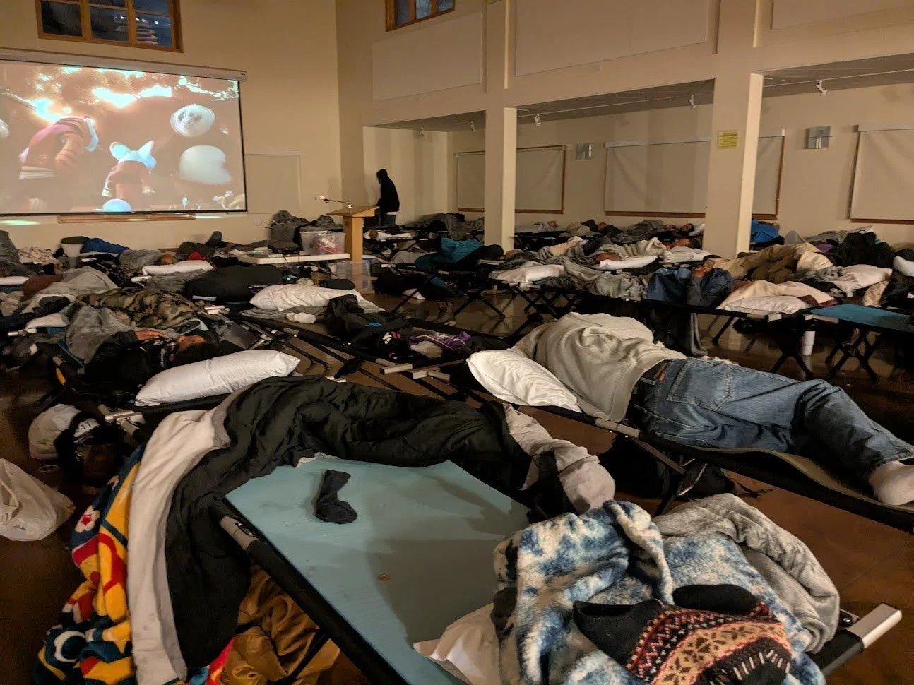 A large room converted into a makeshift shelter with many people sleeping on cots and blankets, some covered with coats or sleeping bags, while others lie directly on the cots.