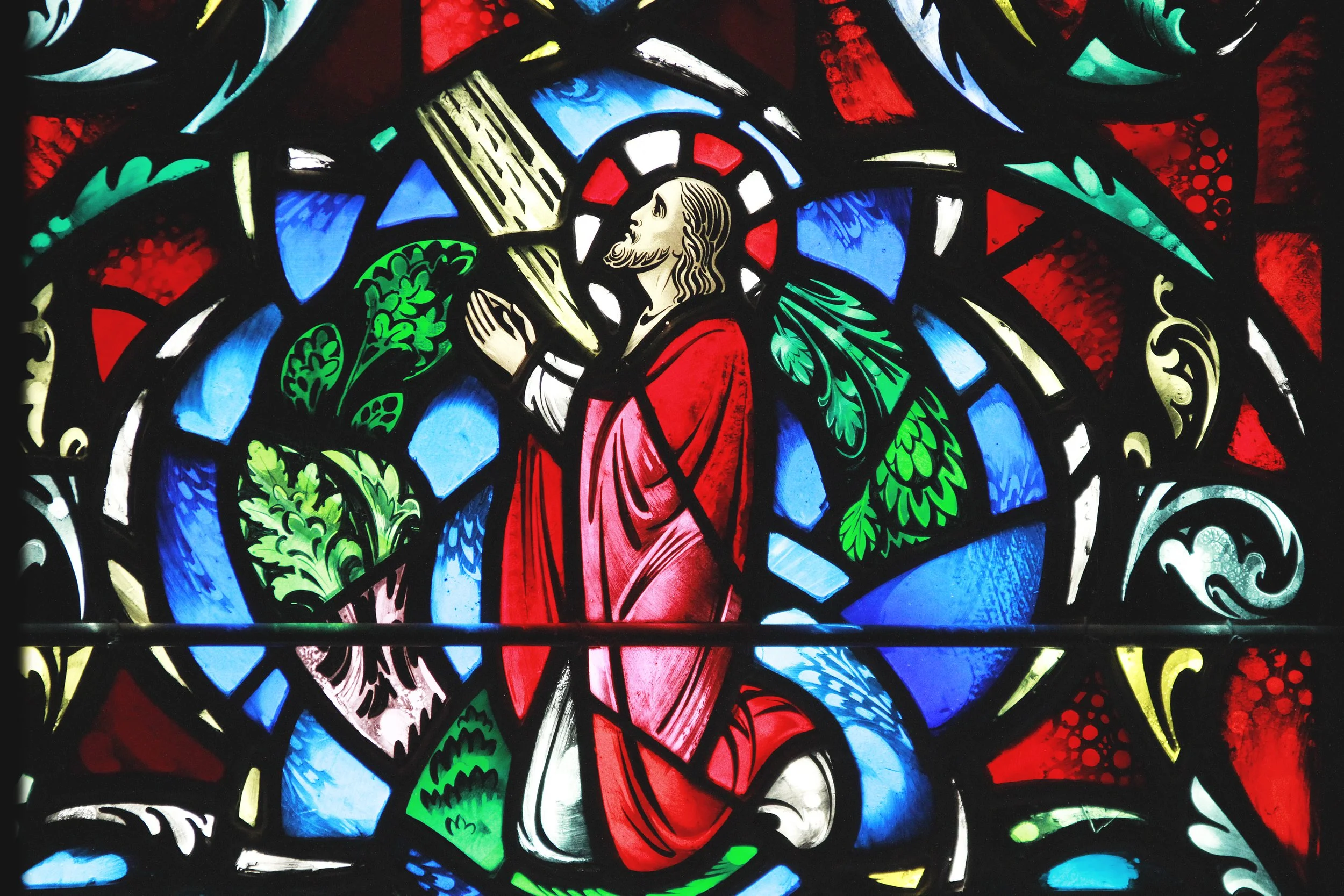 Colorful stained glass window depicting Jesus Christ with a red robe, praying with hands clasped, surrounded by green foliage and abstract patterns in blue, red, yellow, and black.