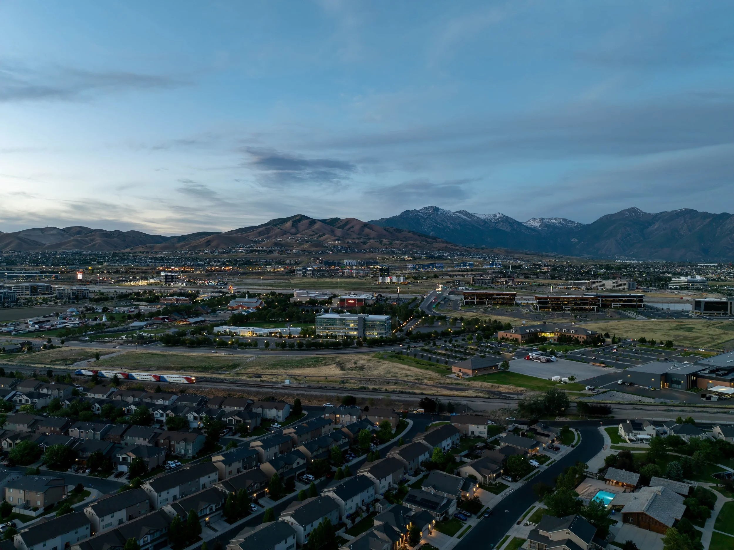 Aerial view of a city with houses in the foreground, commercial buildings and roads in the middle, and mountains in the background during twilight.
