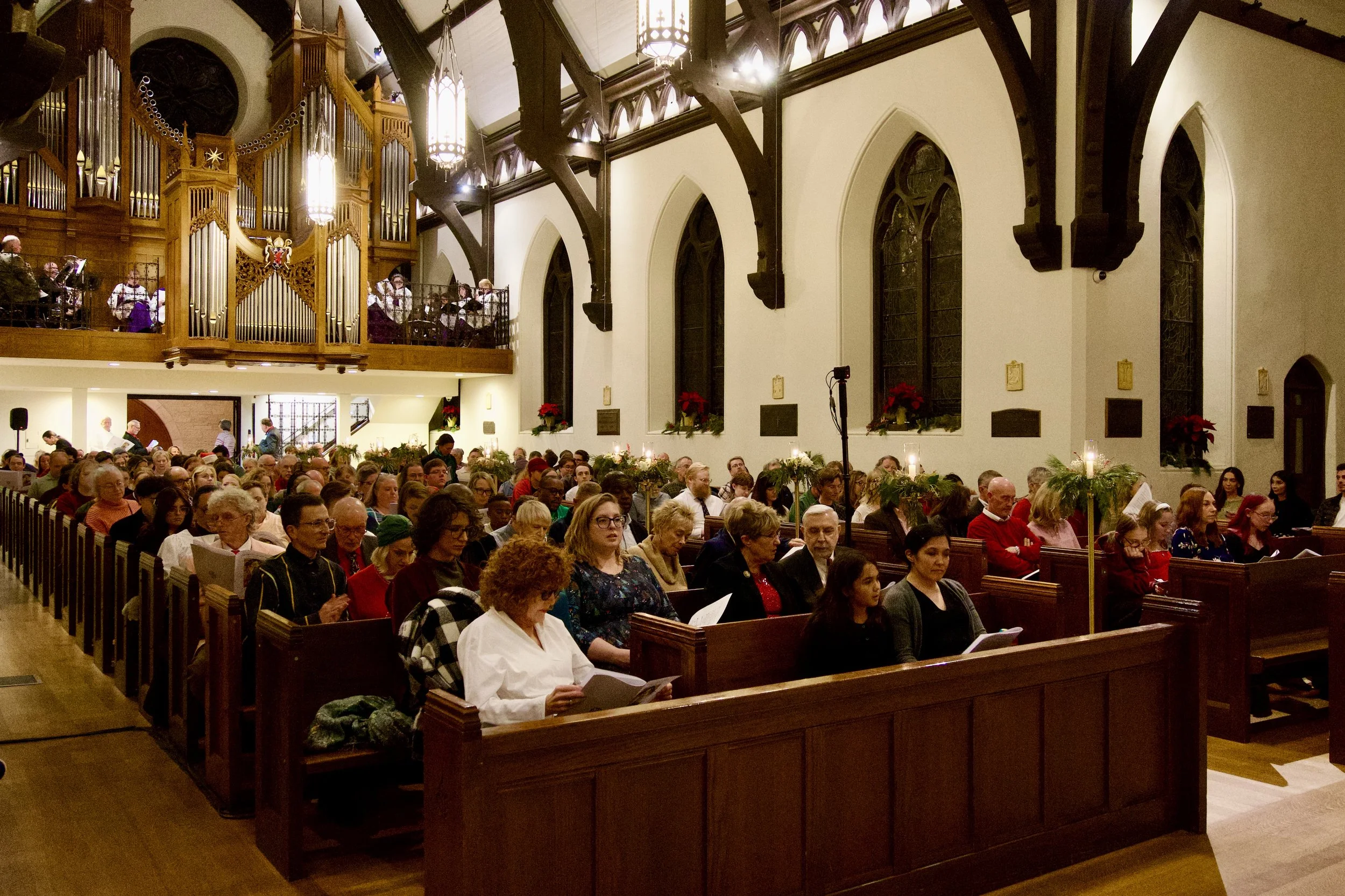 A congregation gathered inside a church during a service, with people seated in wooden pews decorated with Christmas poinsettias and candles. The church has tall stained glass windows and wooden beams, with an organ in the upper balcony.