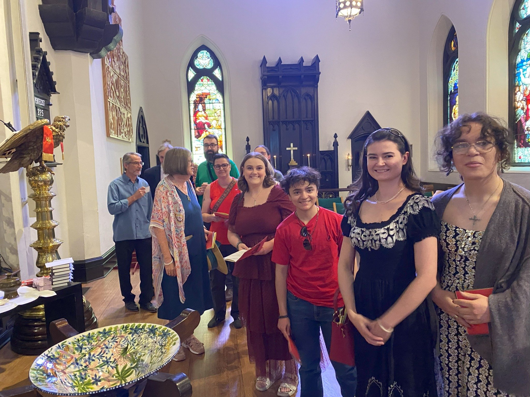 Group of people inside a church, standing in a line and holding hymnals, with stained glass windows and religious decor in the background.