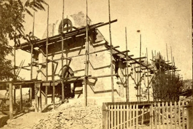 An old black and white photo of the cathedral under construction, surrounded by scaffolding, with trees and a wooden fence nearby.