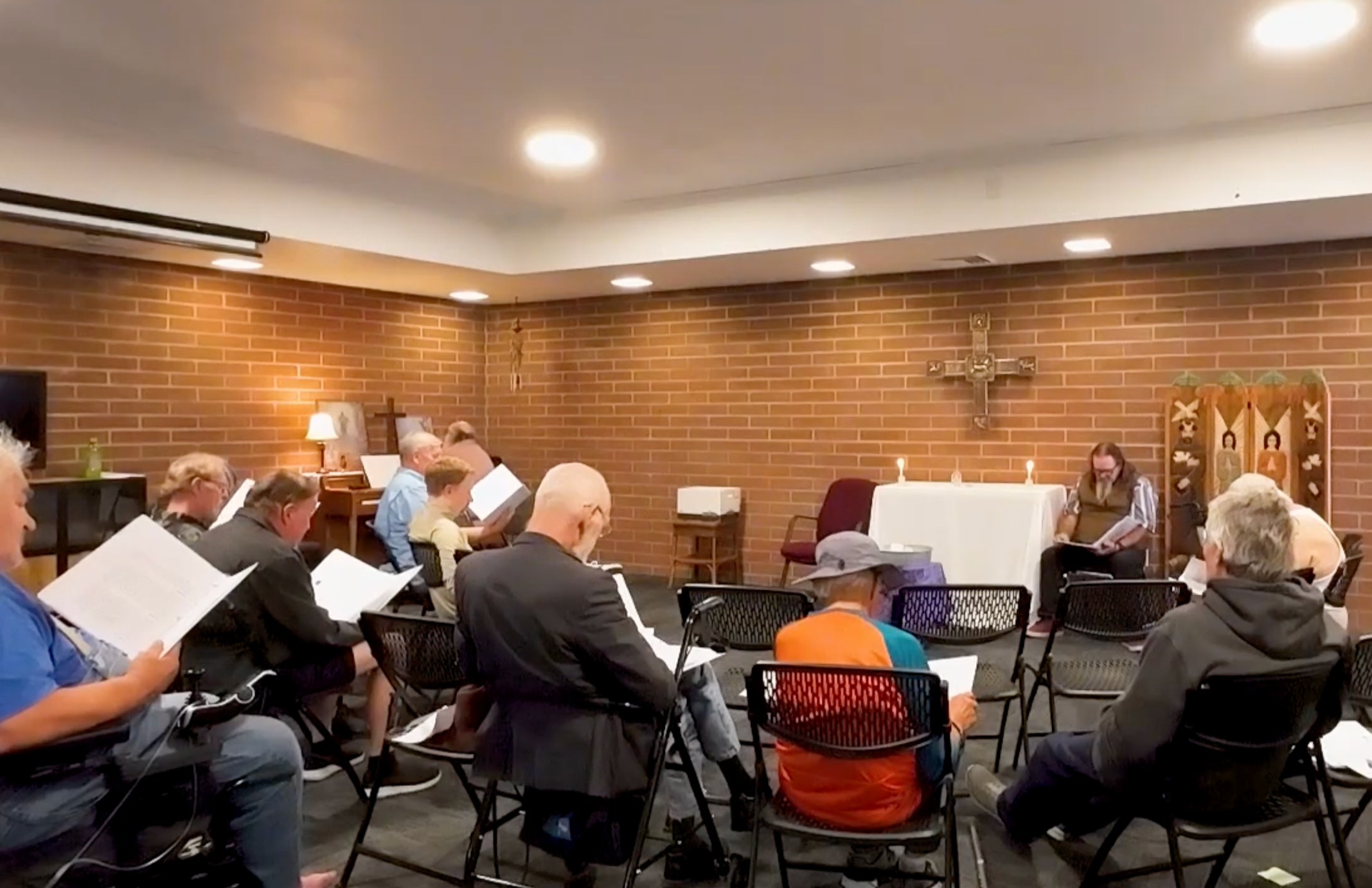 A group of people seated on black chairs in a small church or prayer room, participating in a religious service. There is a cross on the brick wall at the front, with lit candles on a white altar table, and a woman seated to the right near the altar reading from a book.