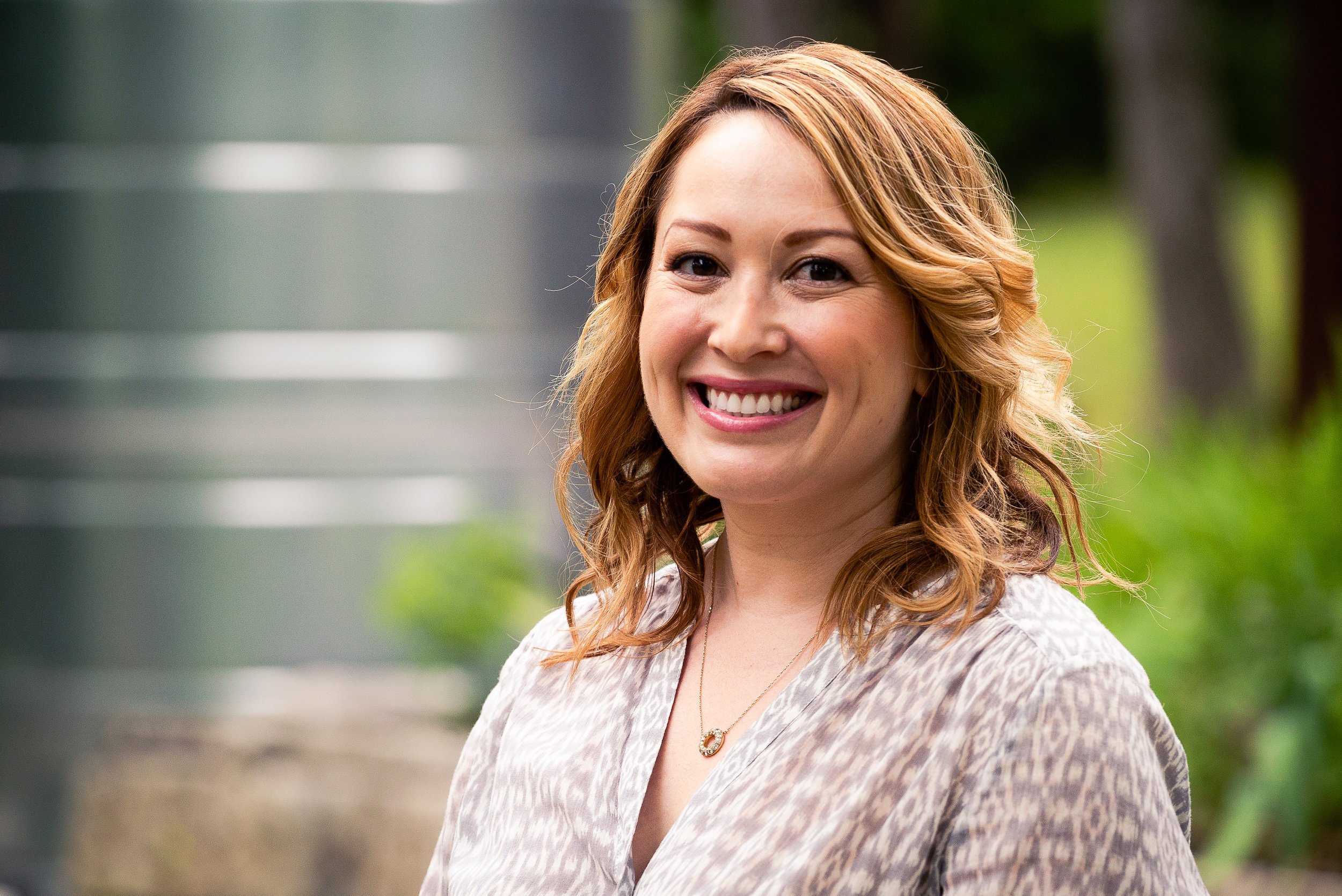A woman with shoulder-length wavy auburn hair smiling outdoors, wearing a light-patterned blouse and a gold necklace with a circular pendant, with greenery in the background.