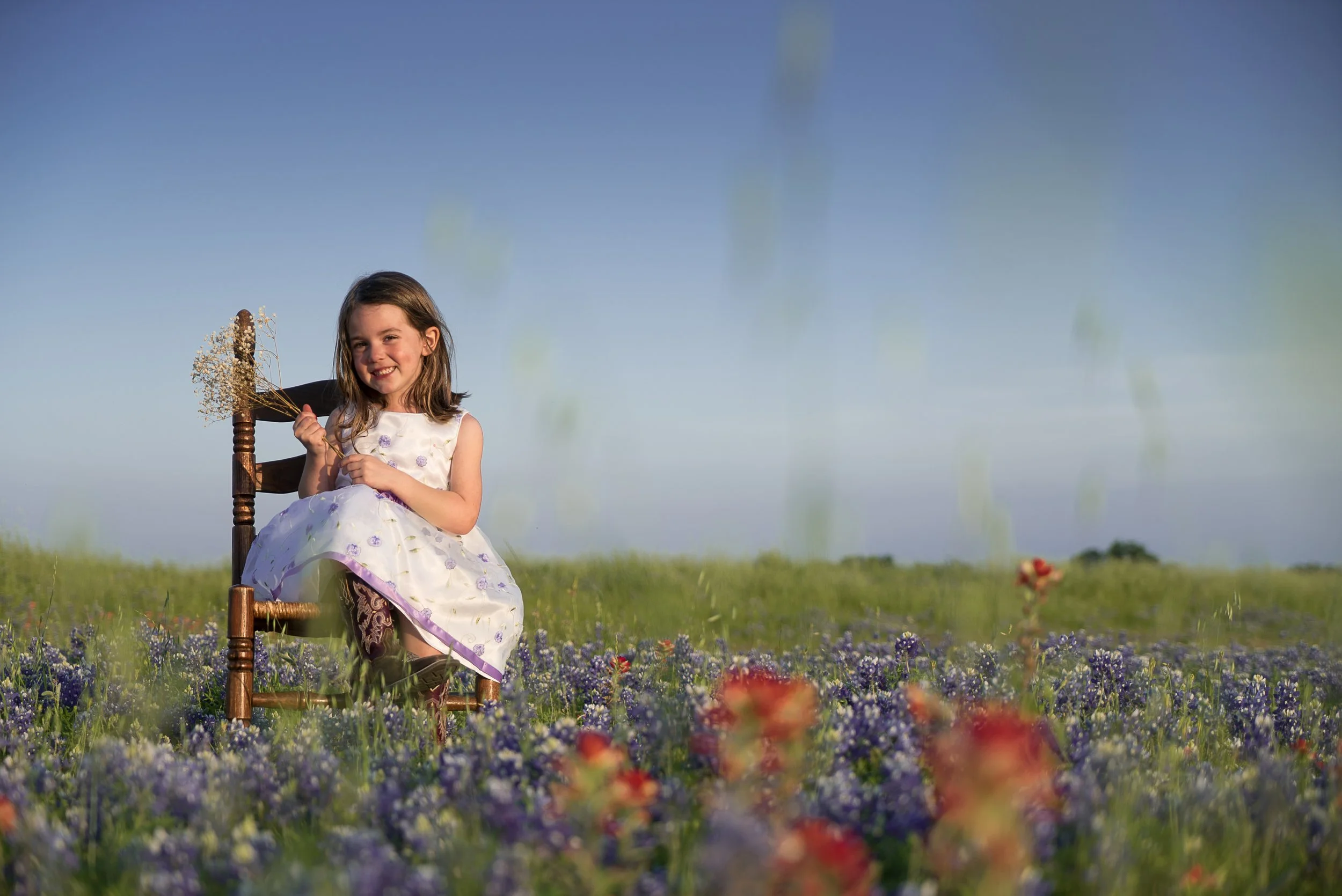 A smiling young girl sitting on a wooden chair in a field of purple flowers, holding a small bouquet of flowers with a clear blue sky in the background.
