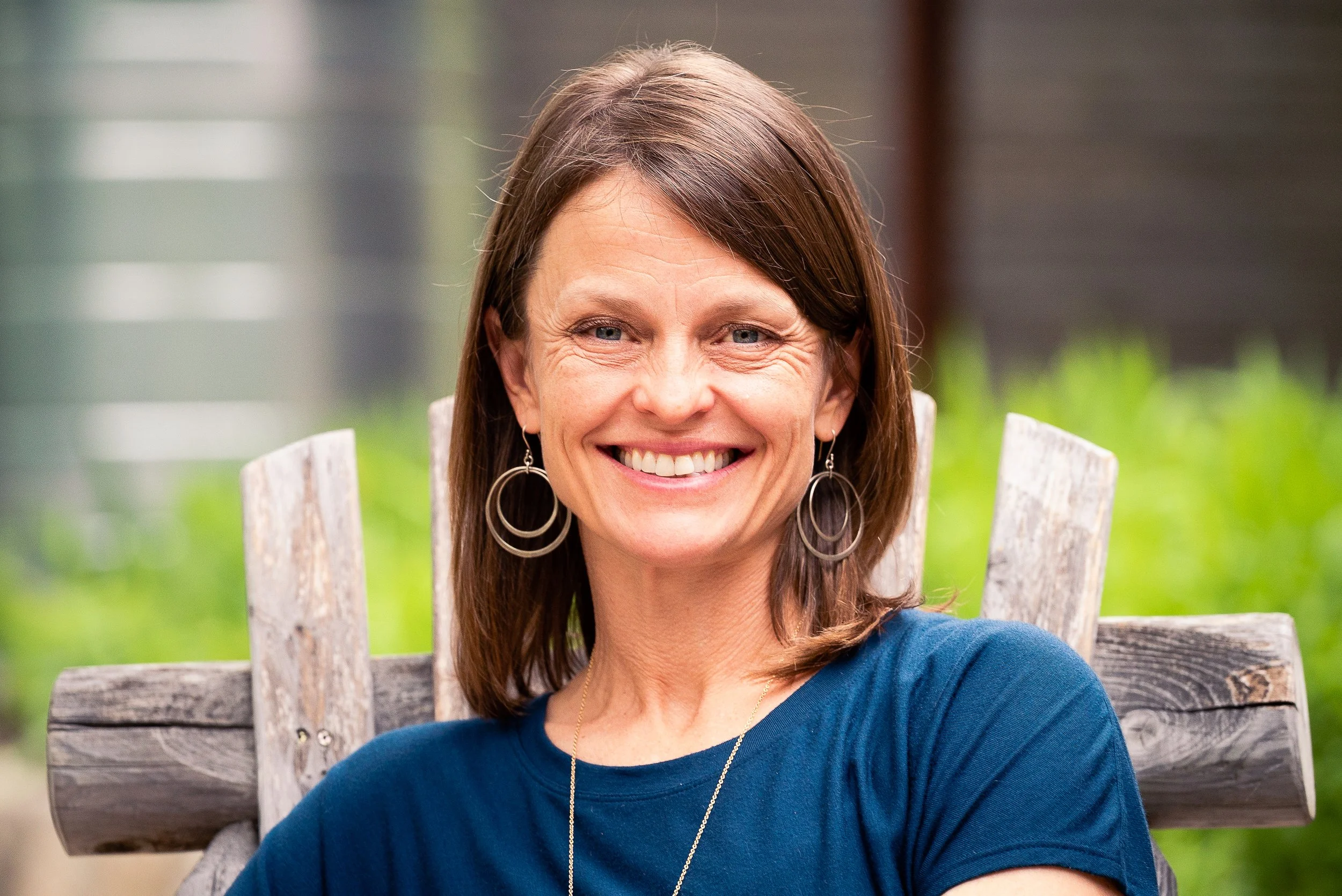 A smiling woman with shoulder-length brown hair, wearing large hoop earrings, a blue shirt, and a necklace, sitting outdoors on a rustic wooden bench with a blurred green background.