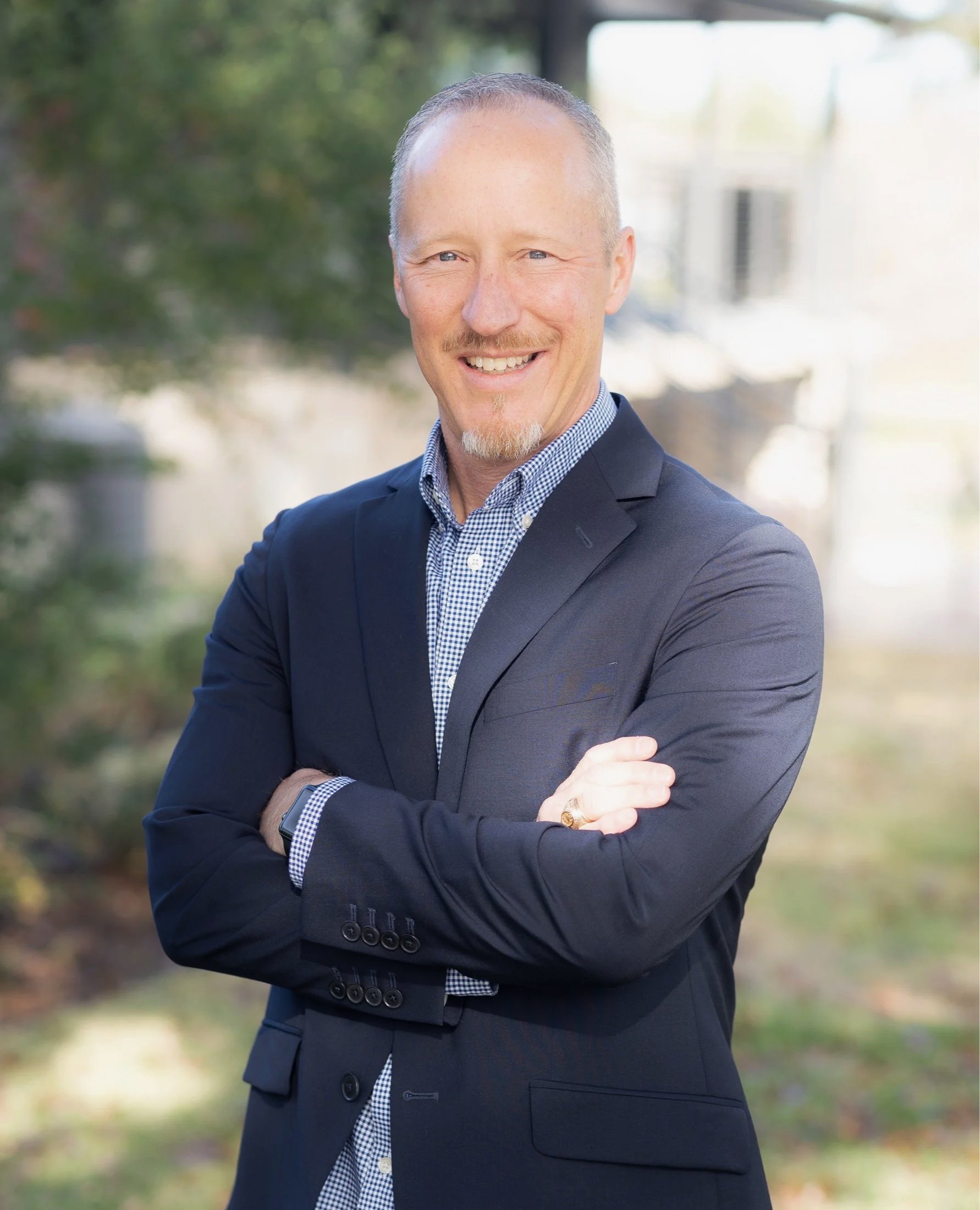 A man smiling, wearing a suit with glasses, standing outdoors with crossed arms.